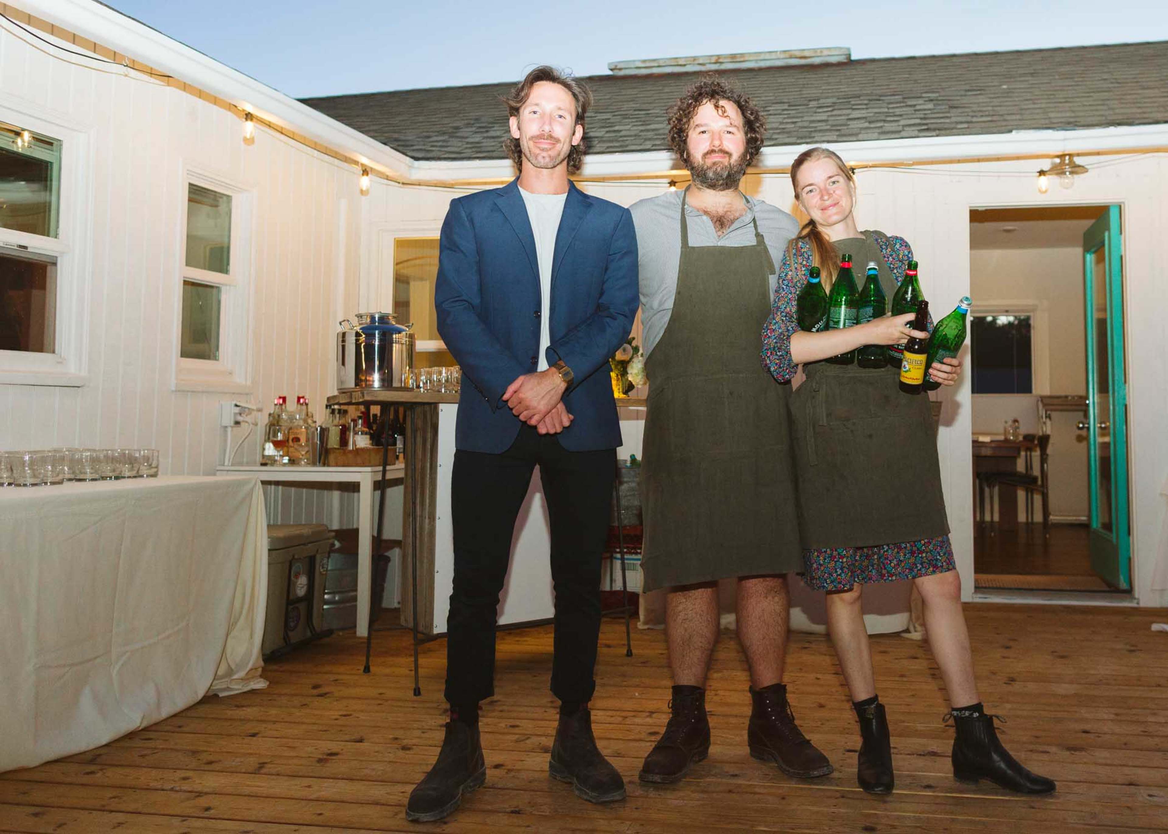 Three individuals stand on a wooden deck, two wearing aprons and holding bottles, while a third is dressed in a blazer.