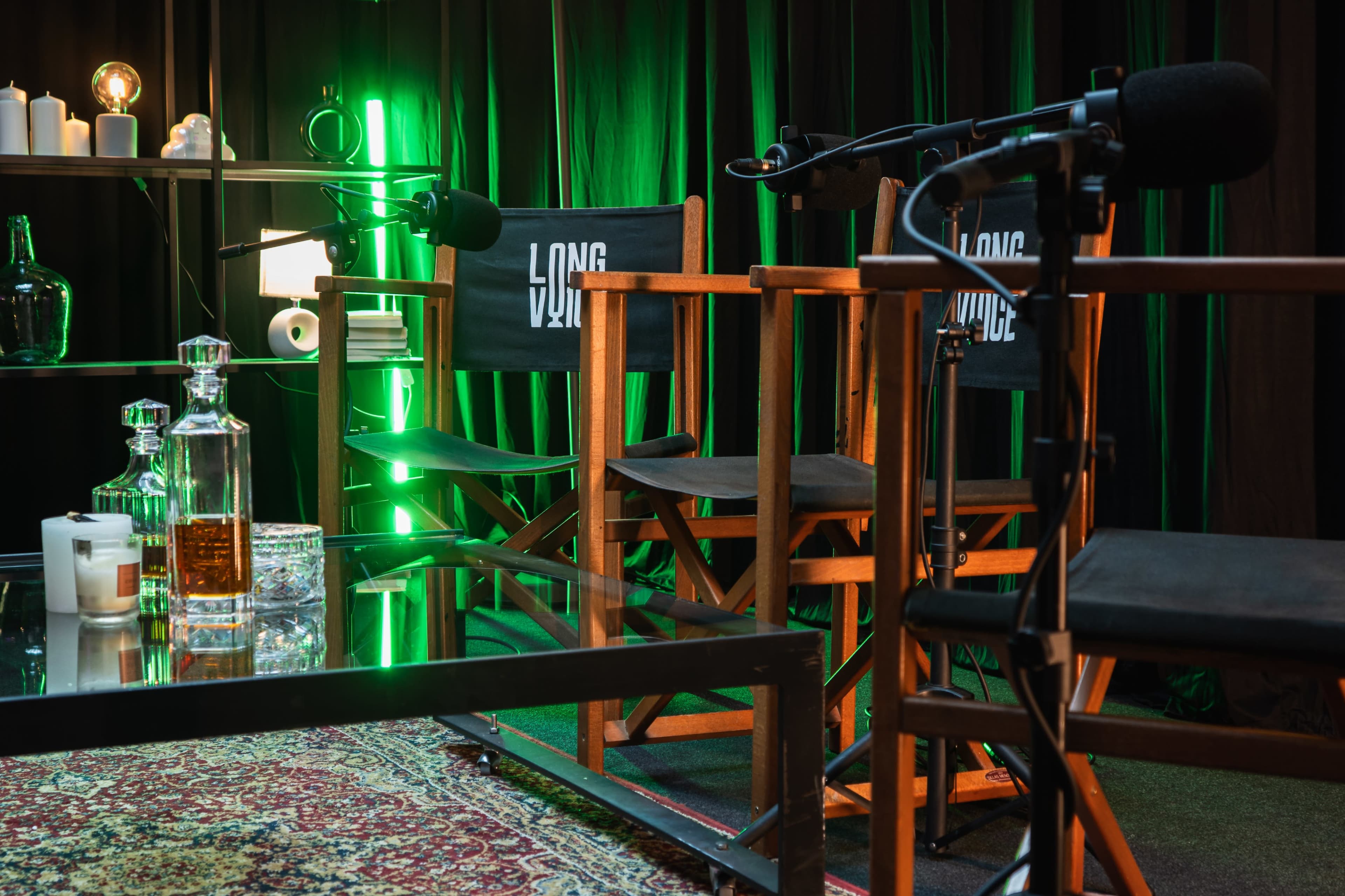 The image shows a set designed for interviews or discussions, featuring three director's chairs, a glass table, decorative bottles, and soft green lighting against a dark backdrop.