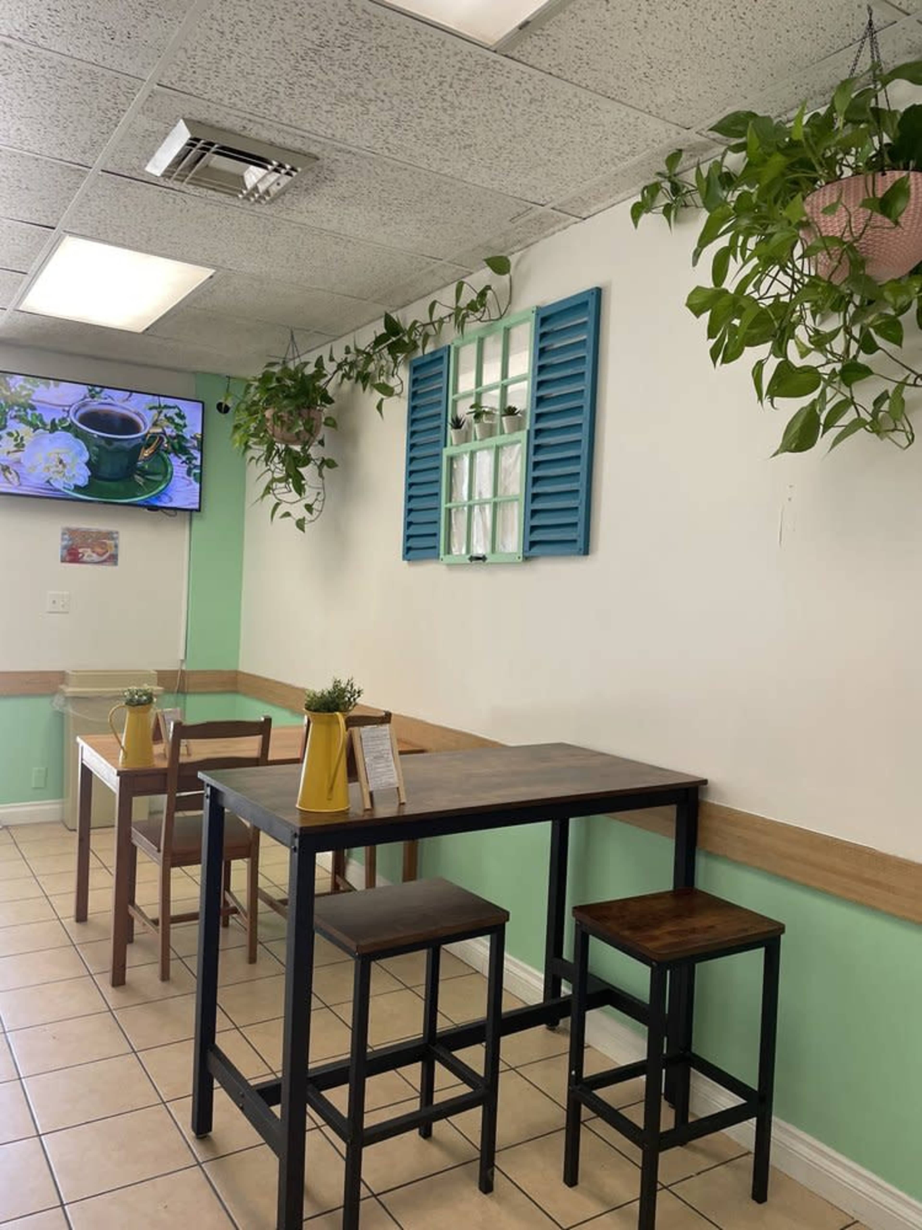The image shows a small cafe interior featuring a table and chairs, green walls, a blue window-shutter decoration, and several hanging plants.