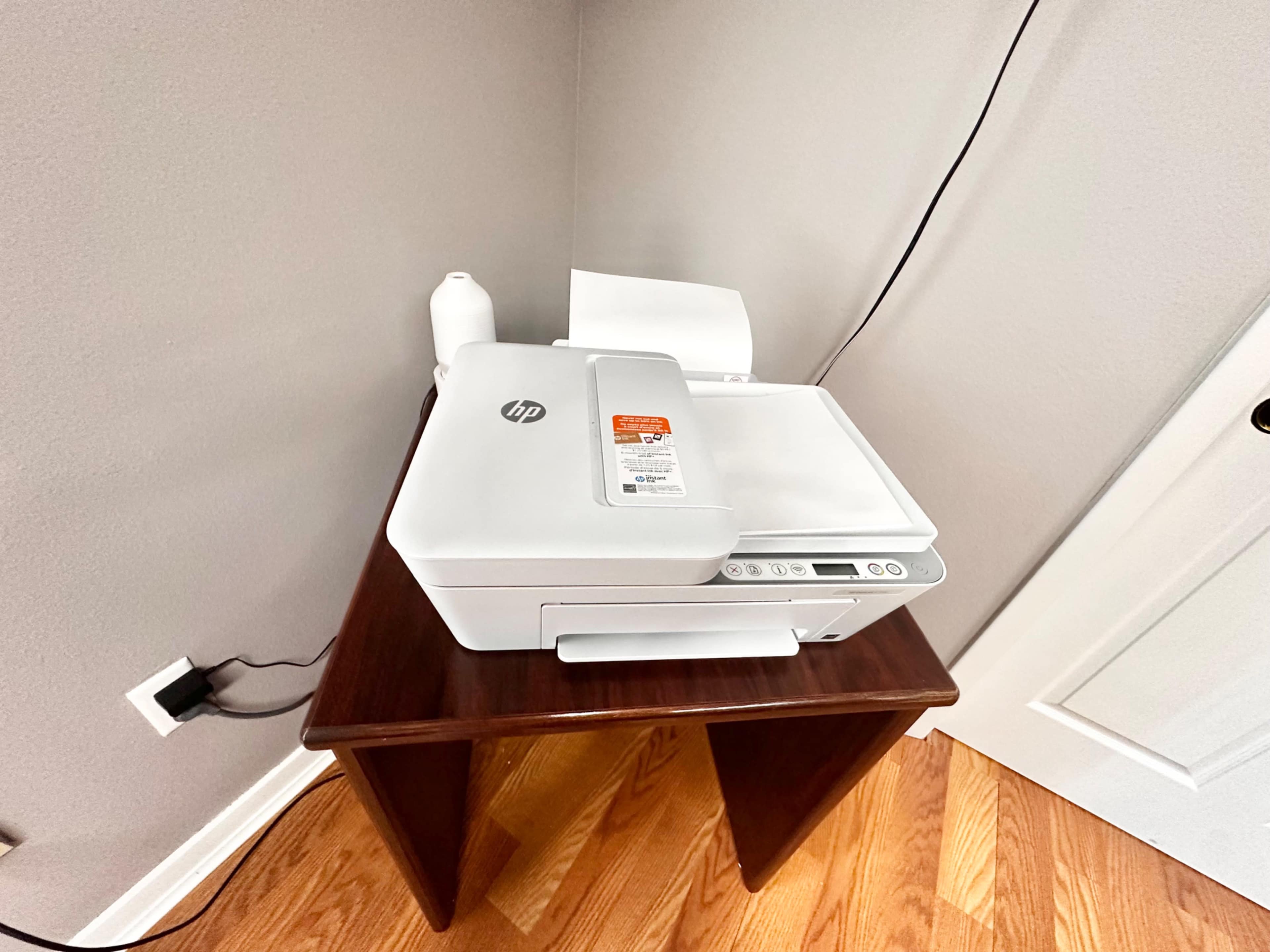 A white HP printer is placed on a brown wooden table against a gray wall, with a sheet of paper partially fed into the printer.