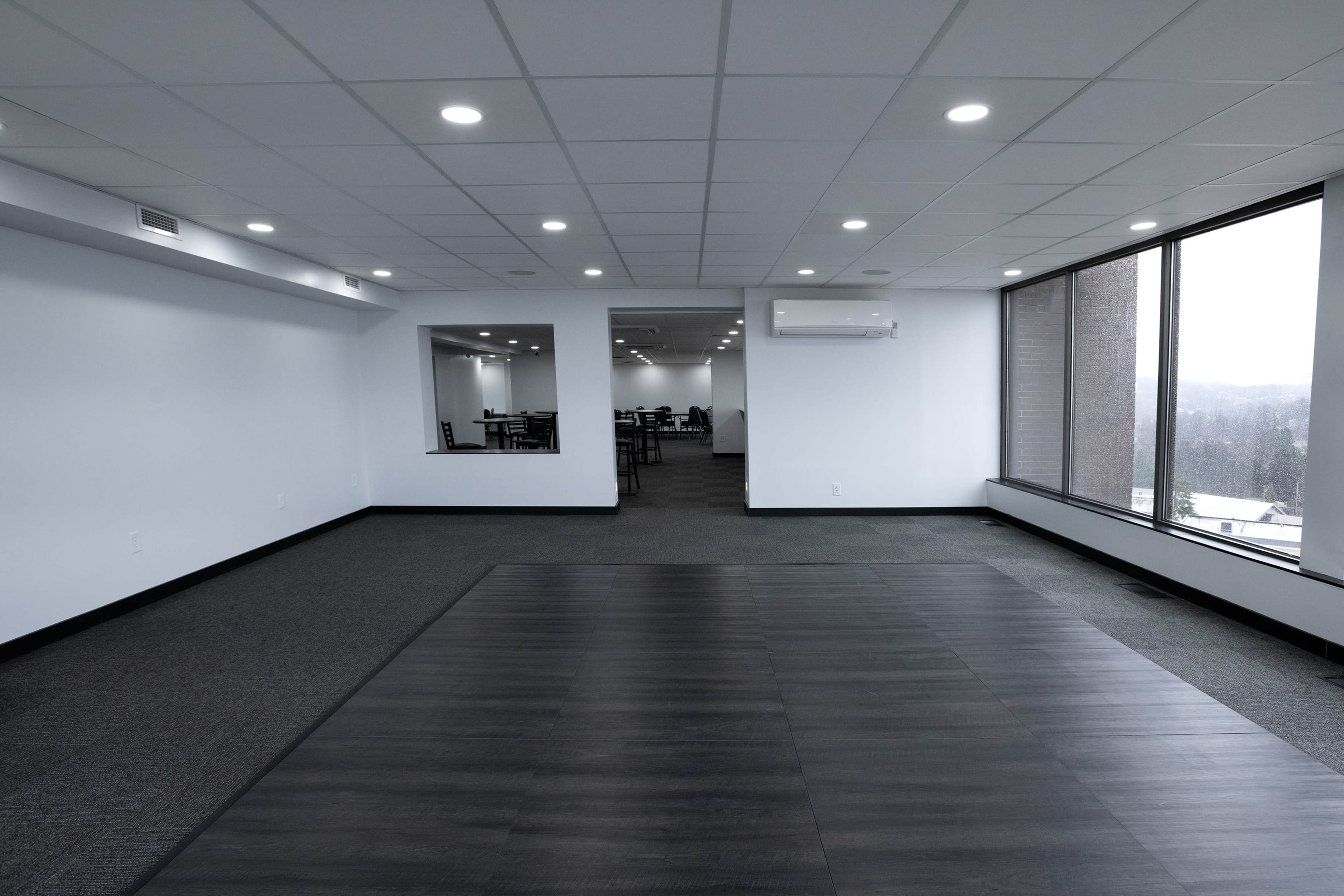 A spacious, empty office area with gray carpet and a large window, revealing a view outside.
