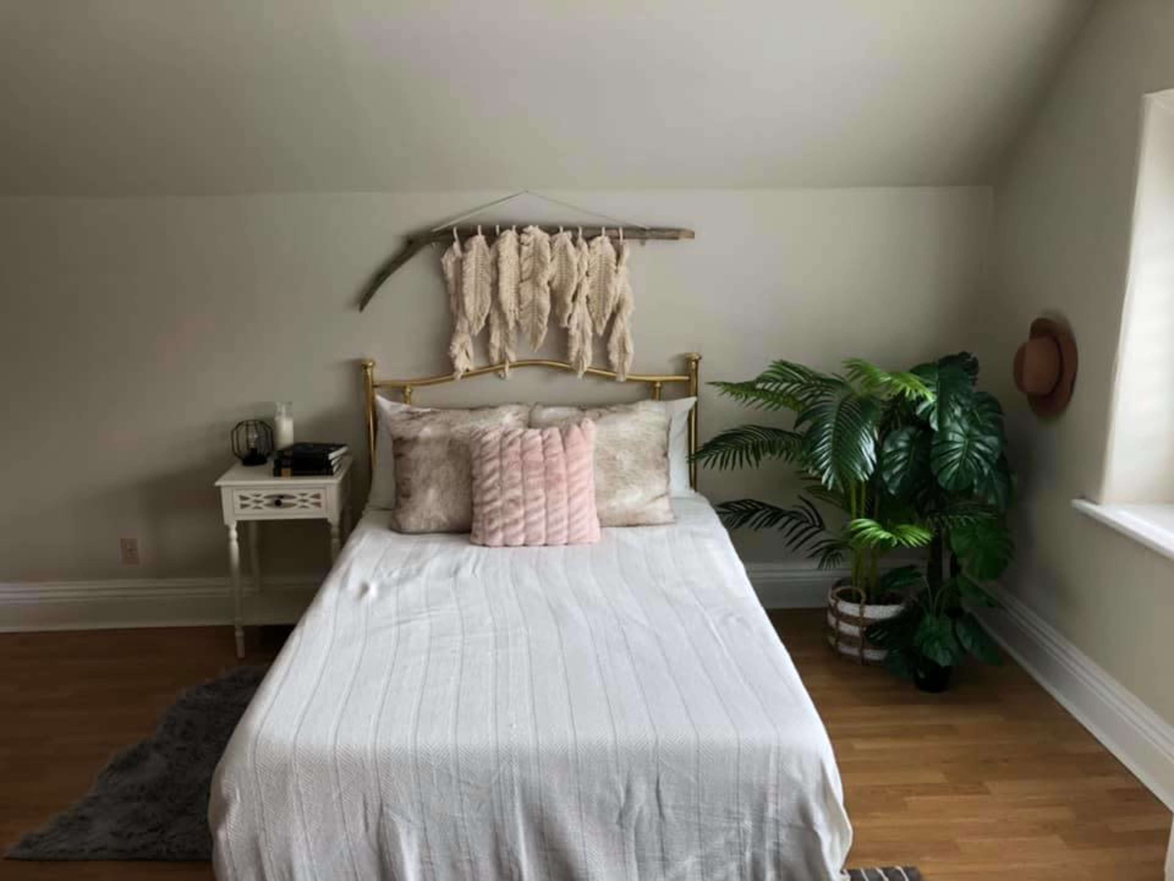 A neatly arranged bedroom features a brass bed with decorative pillows, a nightstand, and a large potted plant against a light-colored wall.