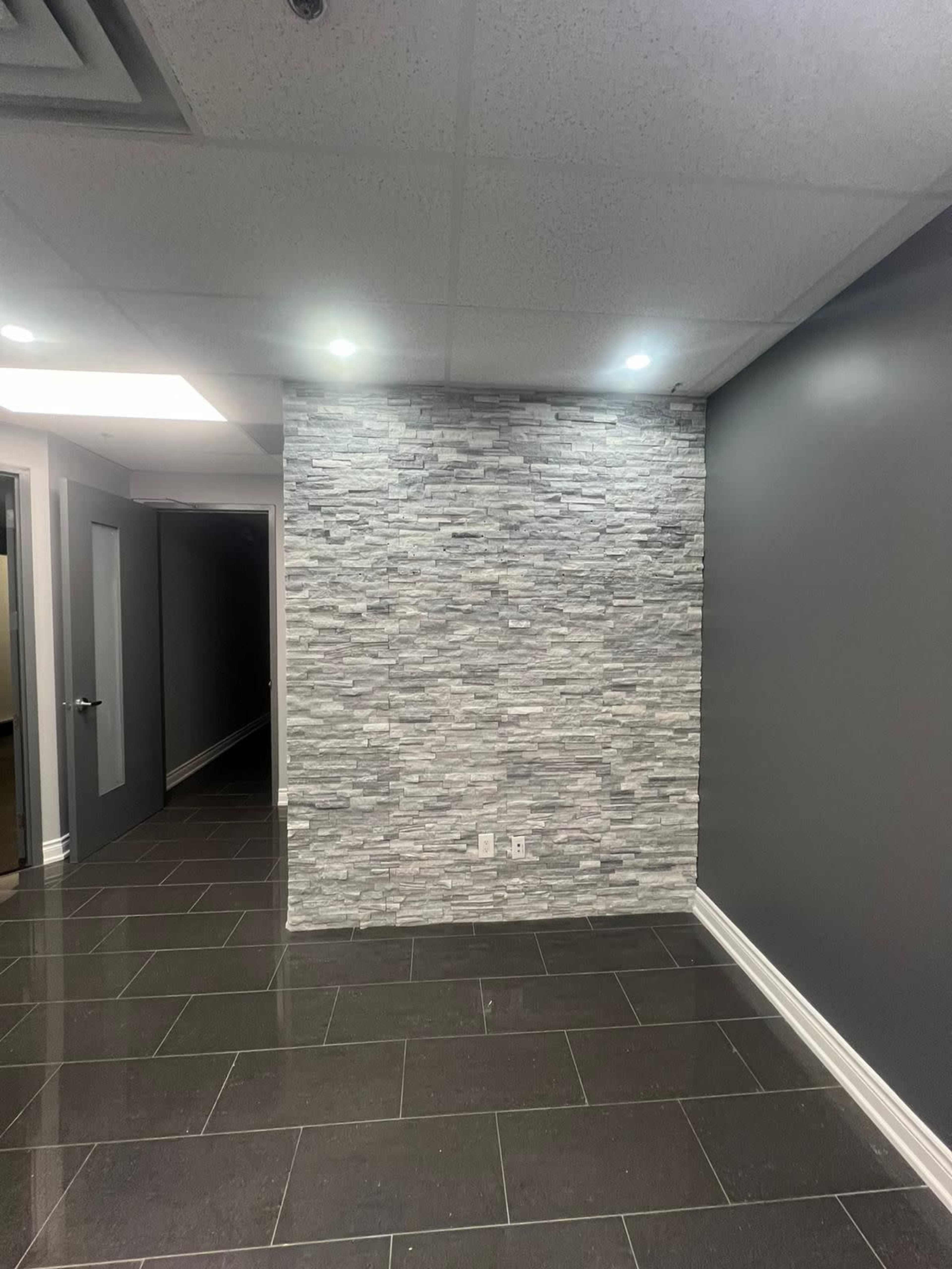 A room with dark floor tiles and a textured stone accent wall, illuminated by recessed lighting, with a doorway visible in the background.