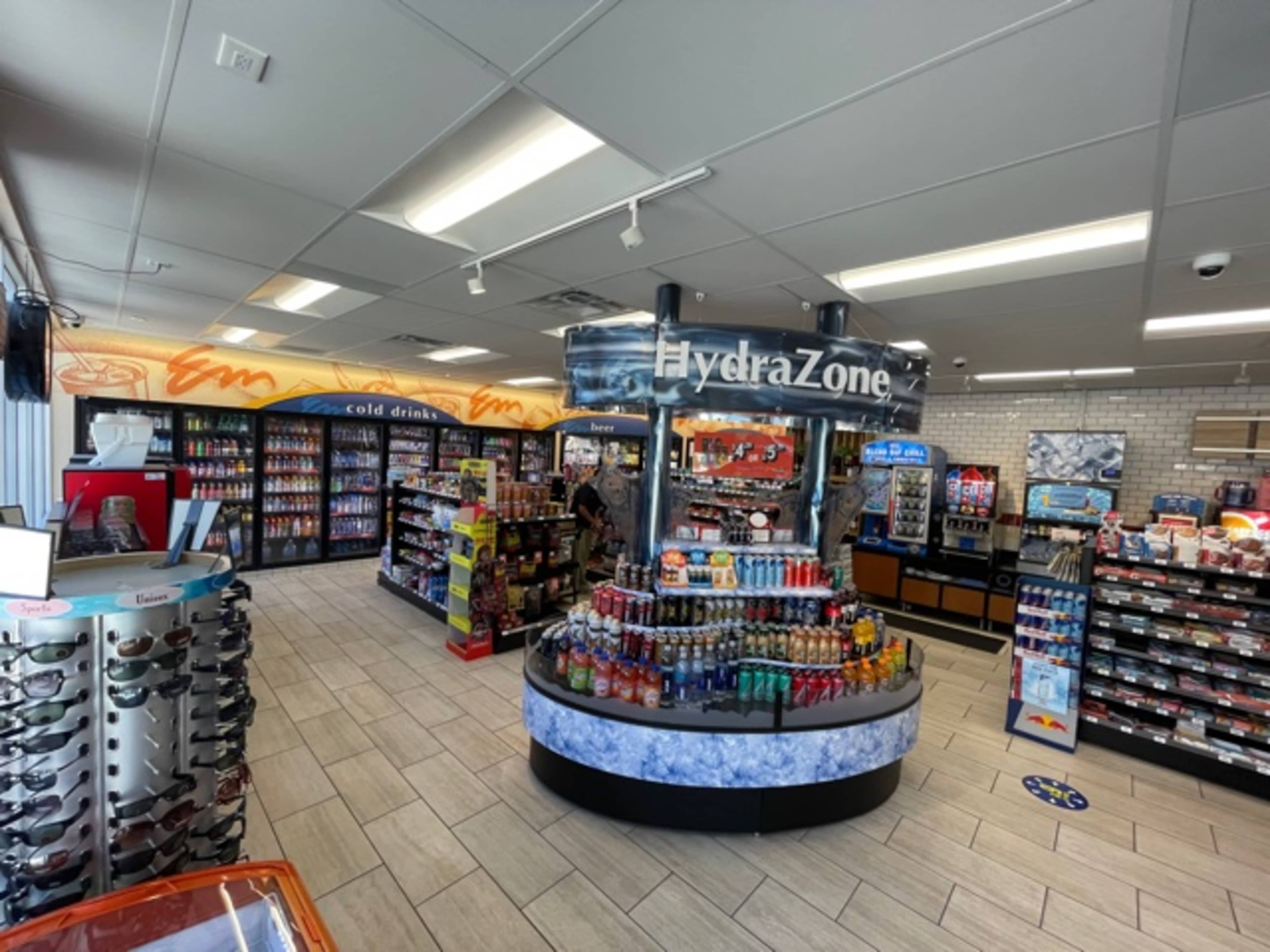 The image shows a convenience store interior featuring a central display for beverages labeled "HydraZone," surrounded by shelves stocked with various drinks and snacks.