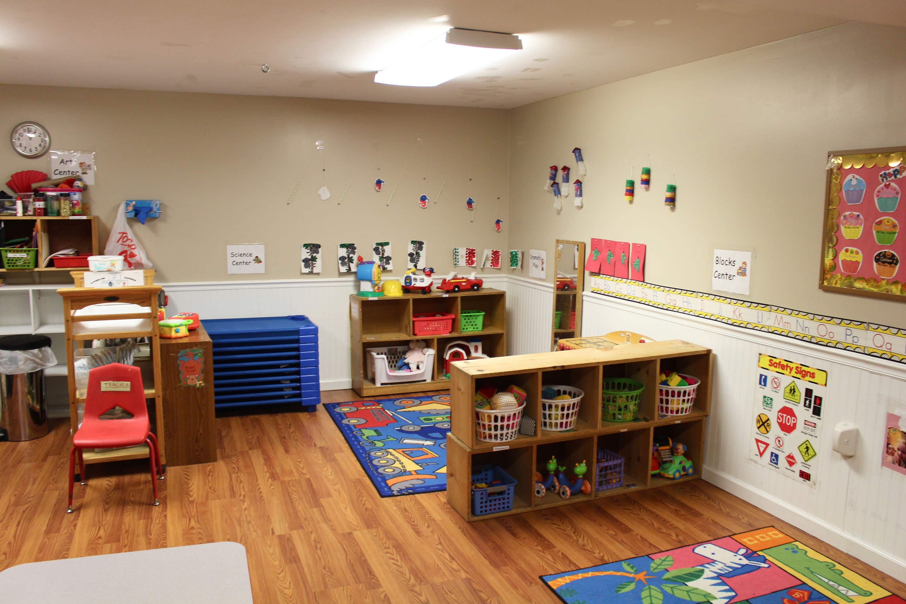 A children's classroom with colorful rugs, storage shelves filled with toys, and labeled areas for different activities.