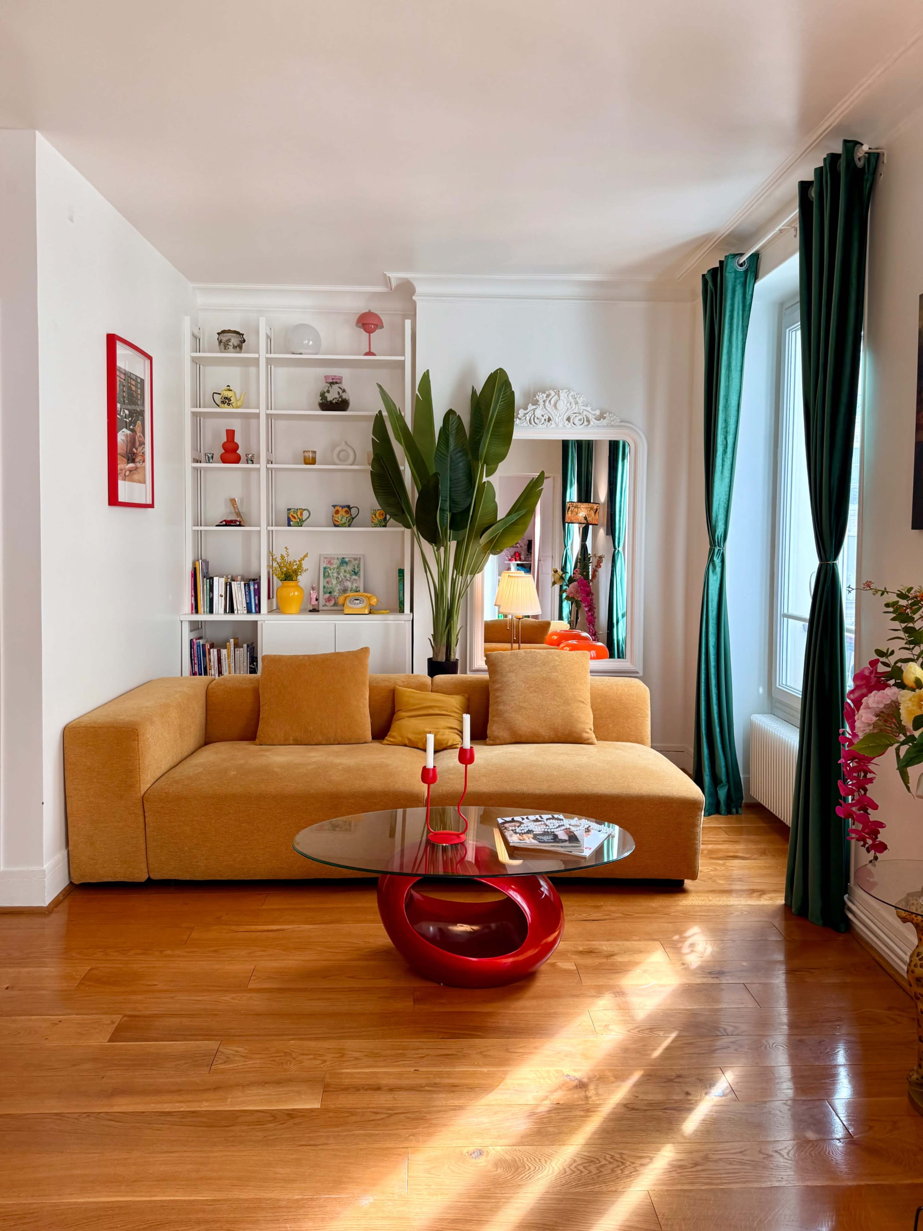 A modern living room features a mustard-yellow sofa, a glass coffee table with red accents, and a tall plant beside a bookshelf filled with various items.