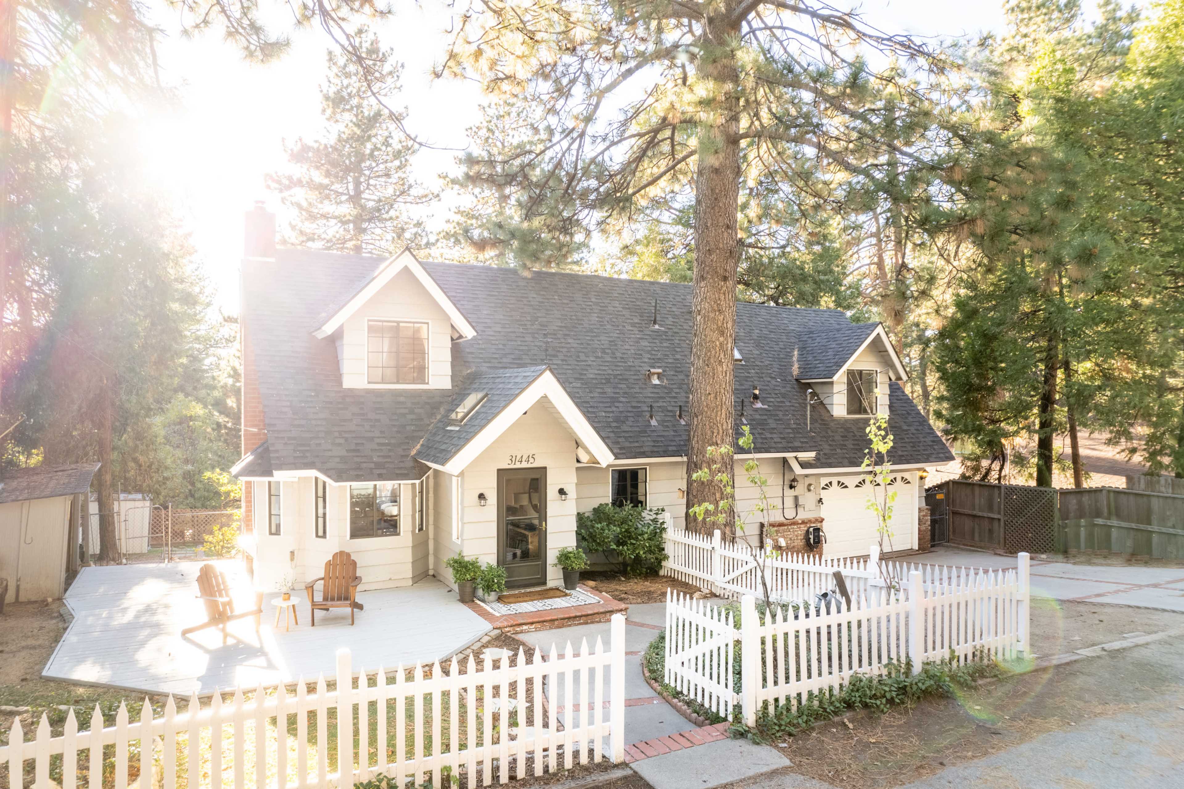 A white house with a dark roof, surrounded by trees, features a front porch with Adirondack chairs and a white picket fence.