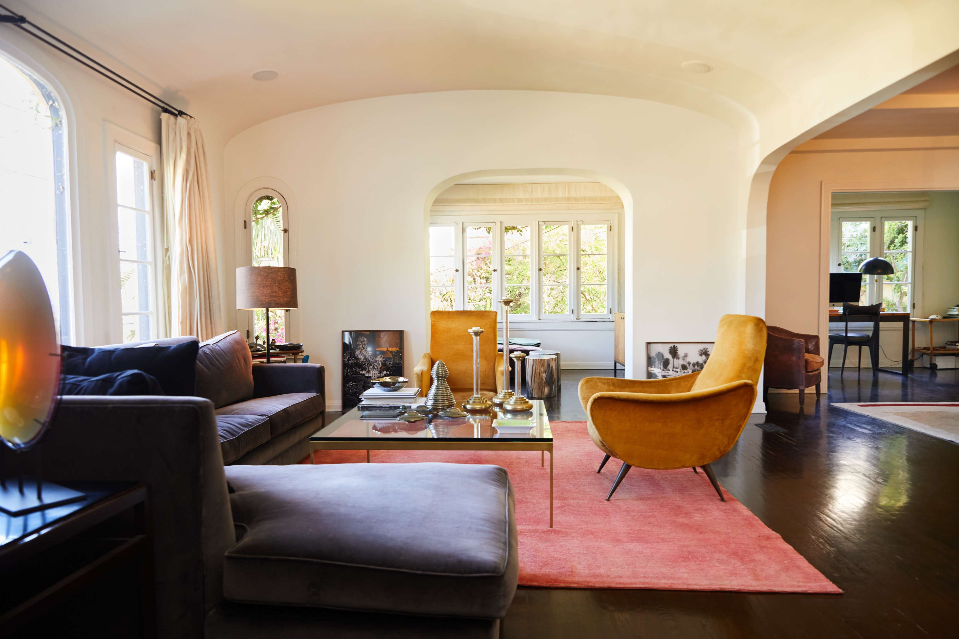A modern living room with a gray sectional sofa, an orange armchair, a glass coffee table, and large windows allowing natural light to fill the space.