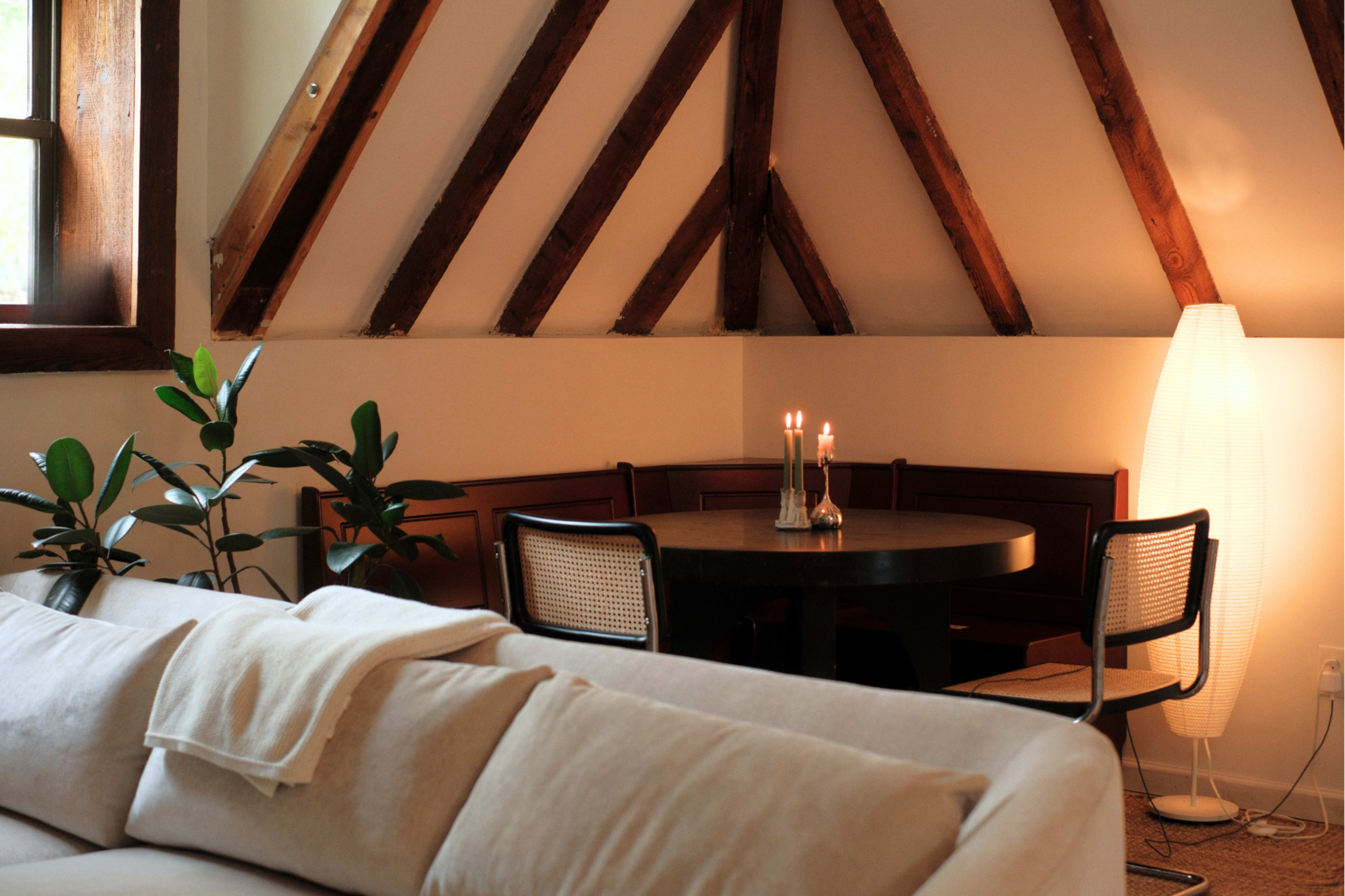 The image shows a cozy interior featuring a round dining table with candles, surrounded by a curved bench and modern chairs, beneath a ceiling with exposed wooden beams.