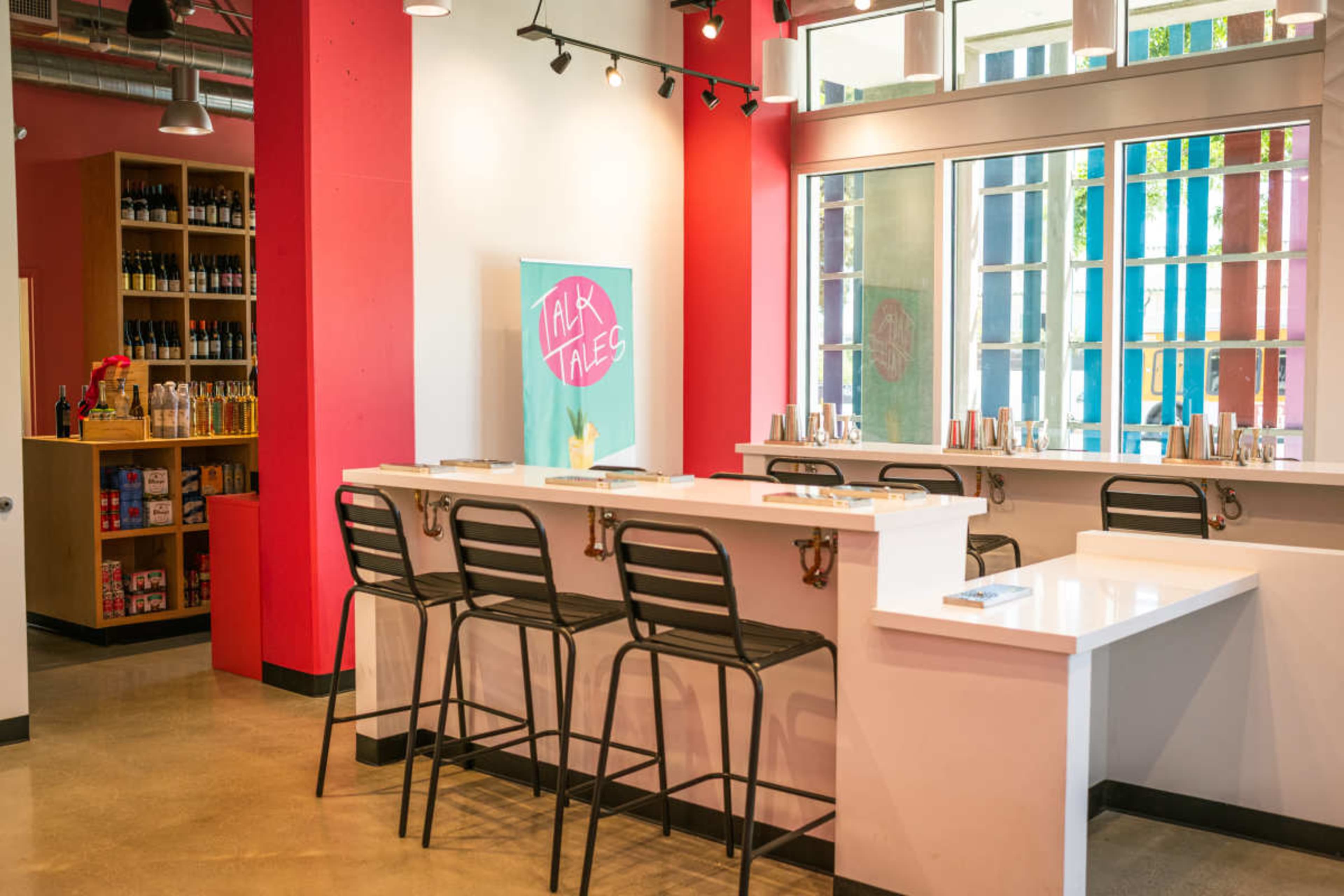 The image shows a modern tasting room with a white countertop bar, black stools, and large windows, set against red and beige walls adorned with shelves of bottles.