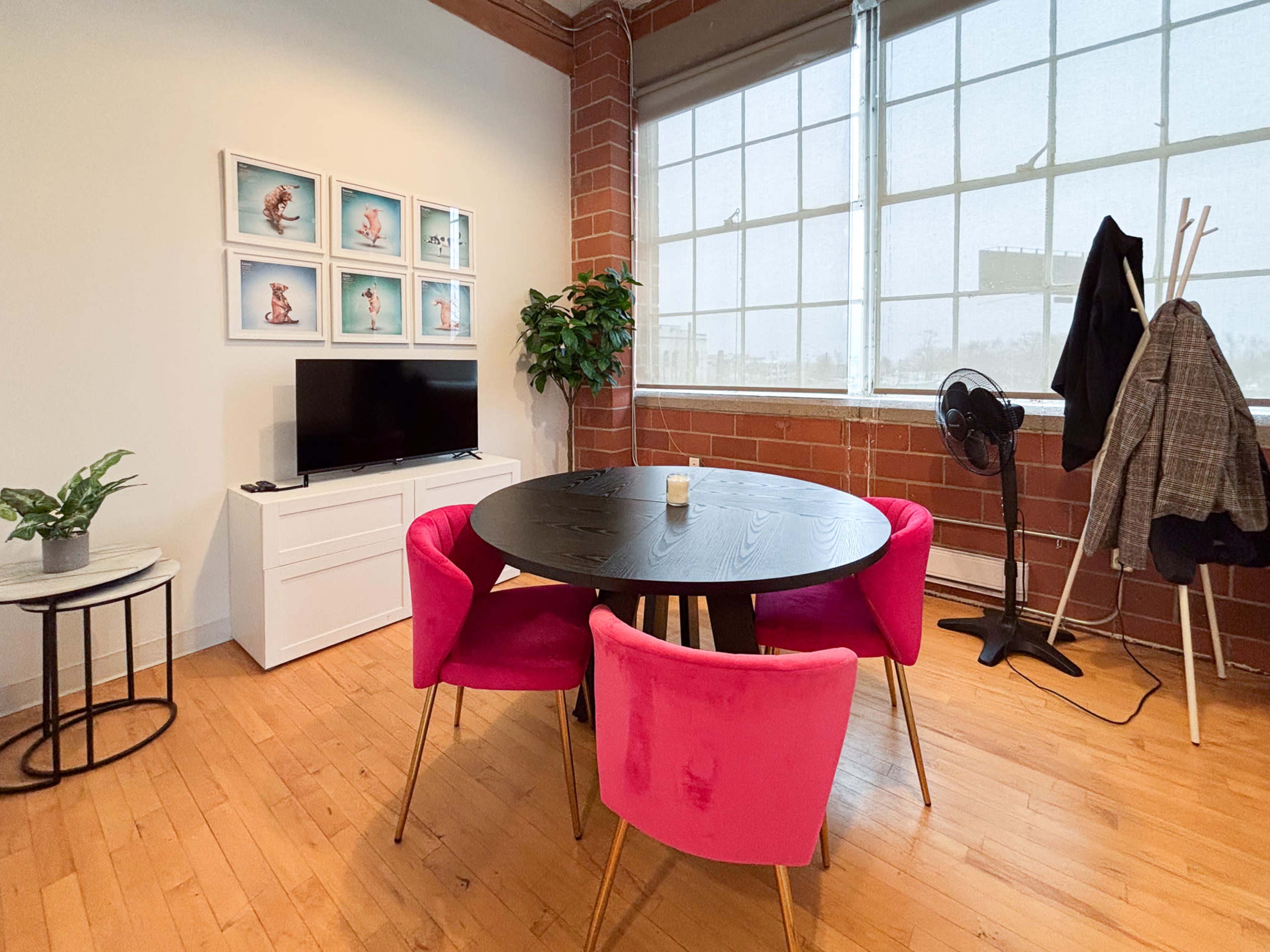 A dining area features a round black table surrounded by pink chairs, with a television on a white cabinet and large windows providing natural light.