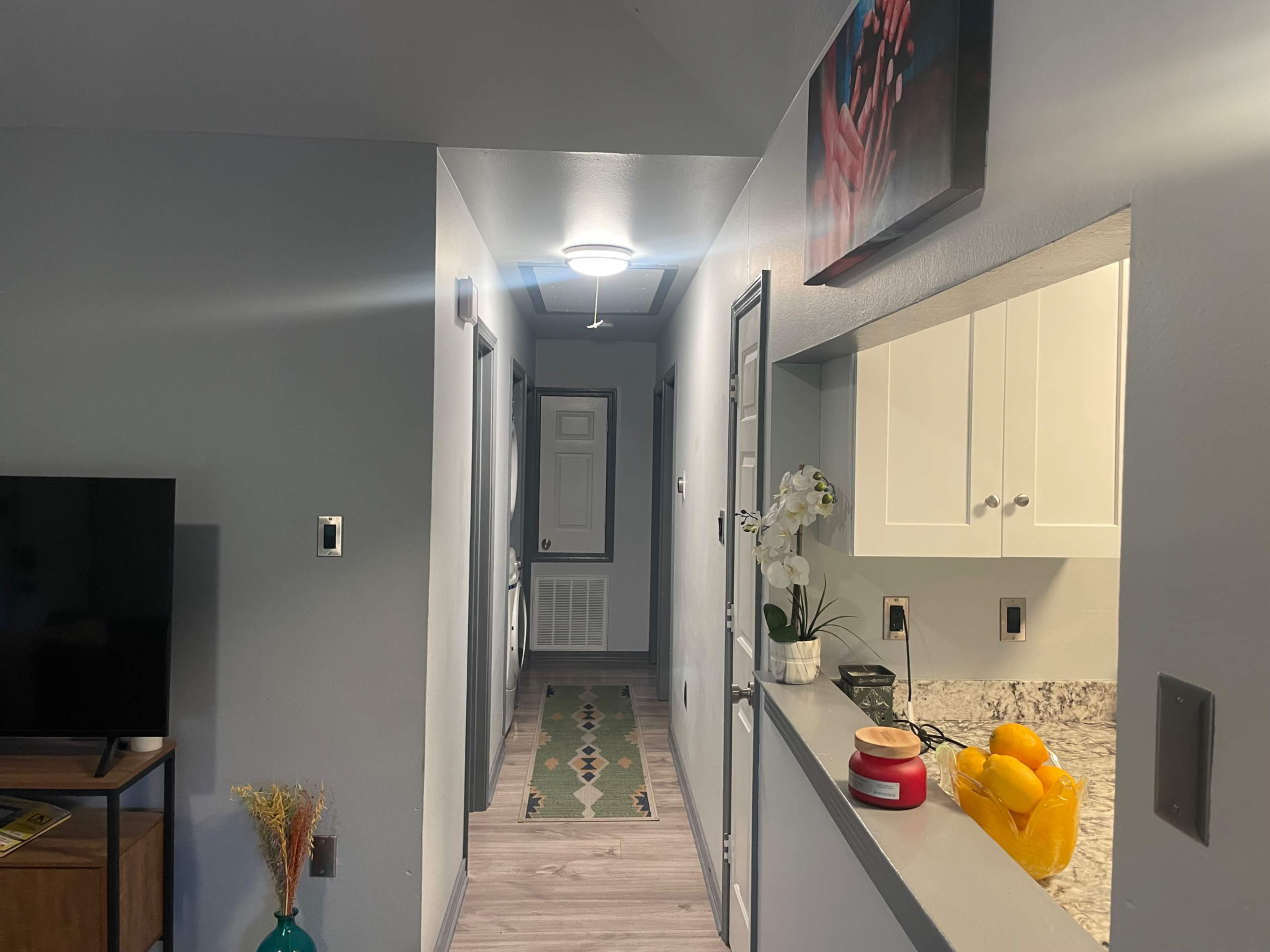 The image shows a modern hallway leading to a door, with a kitchen area visible on the right and a television in the living area.