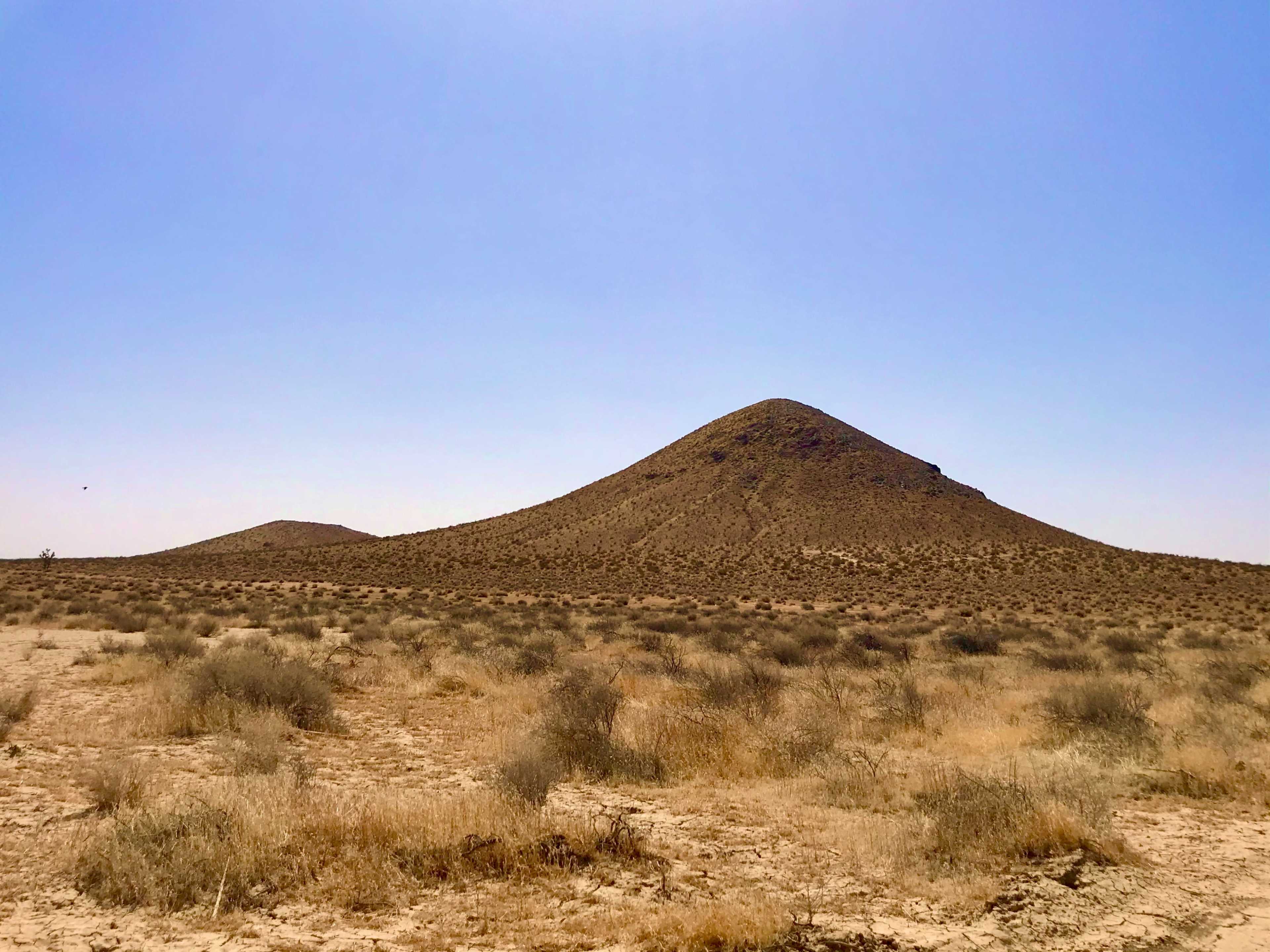 A barren landscape features a prominent conical hill surrounded by sparse vegetation under a clear blue sky.