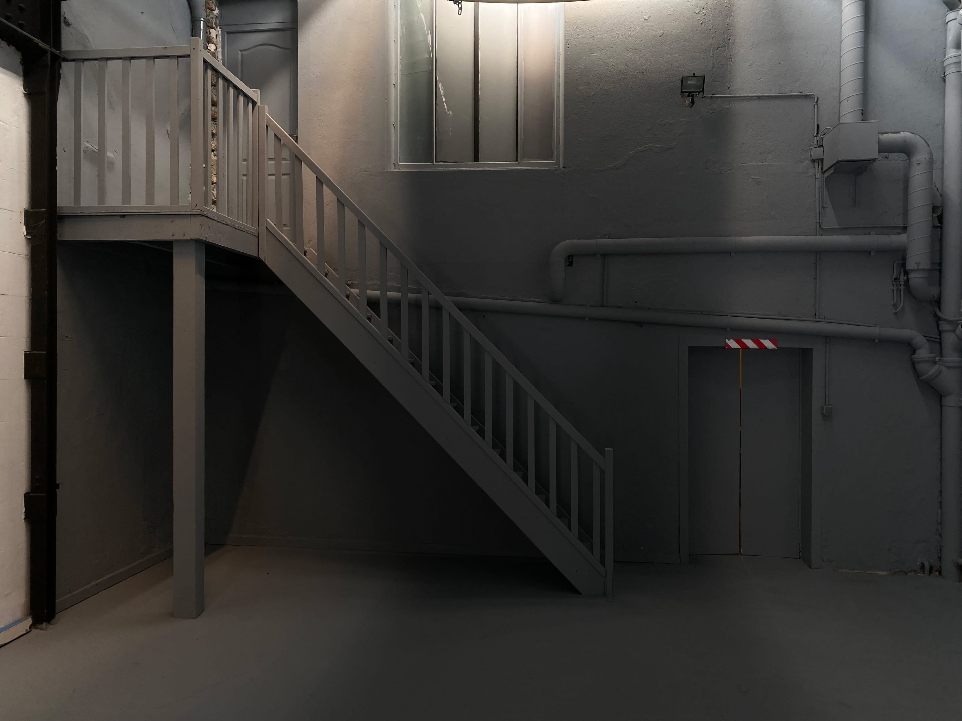 A gray staircase leads to a doorway on the upper level of an industrial-style room with exposed pipes and minimal lighting.