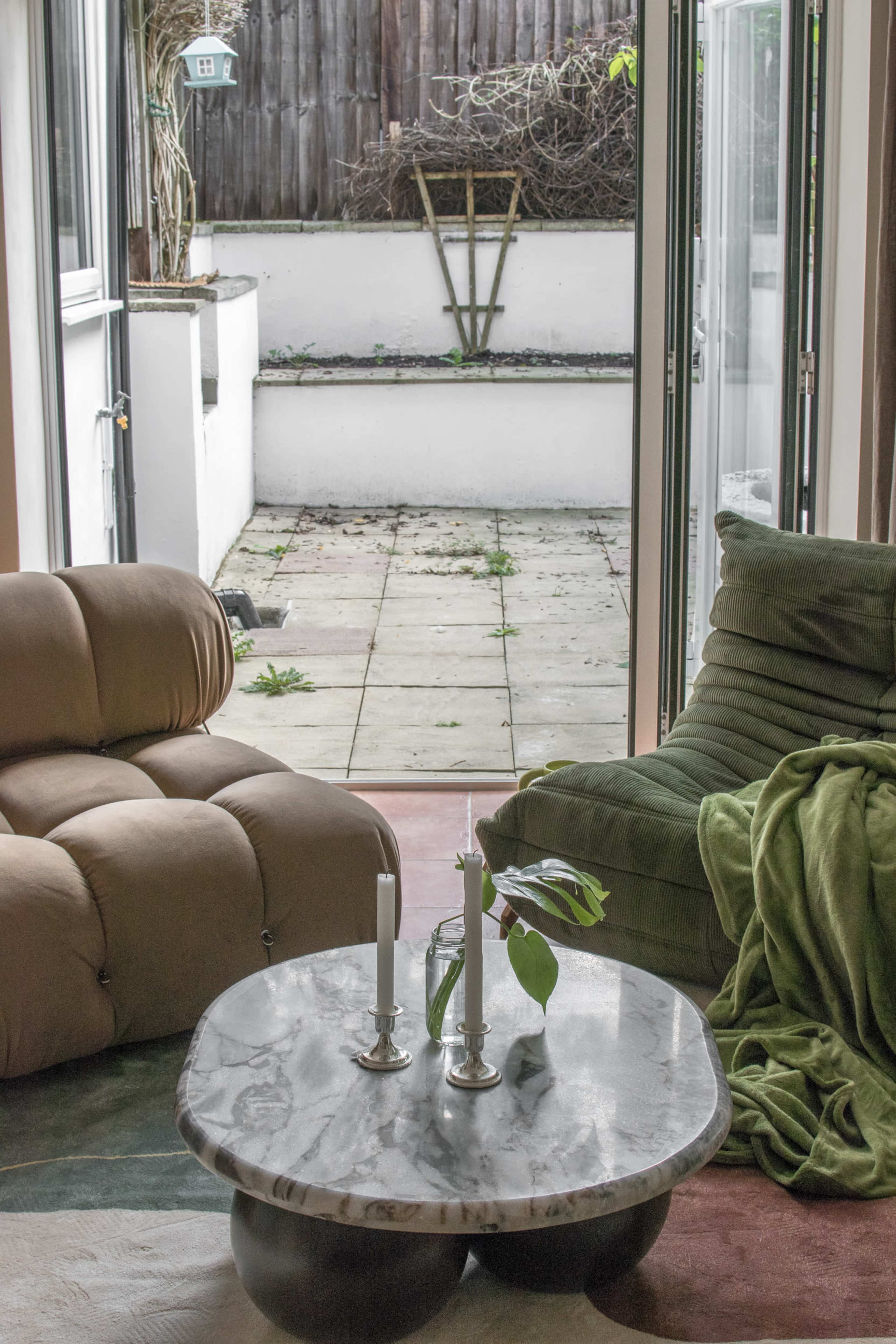 A cozy living area features two plush chairs flanking a round marble coffee table with two candles, overlooking a small outdoor patio.