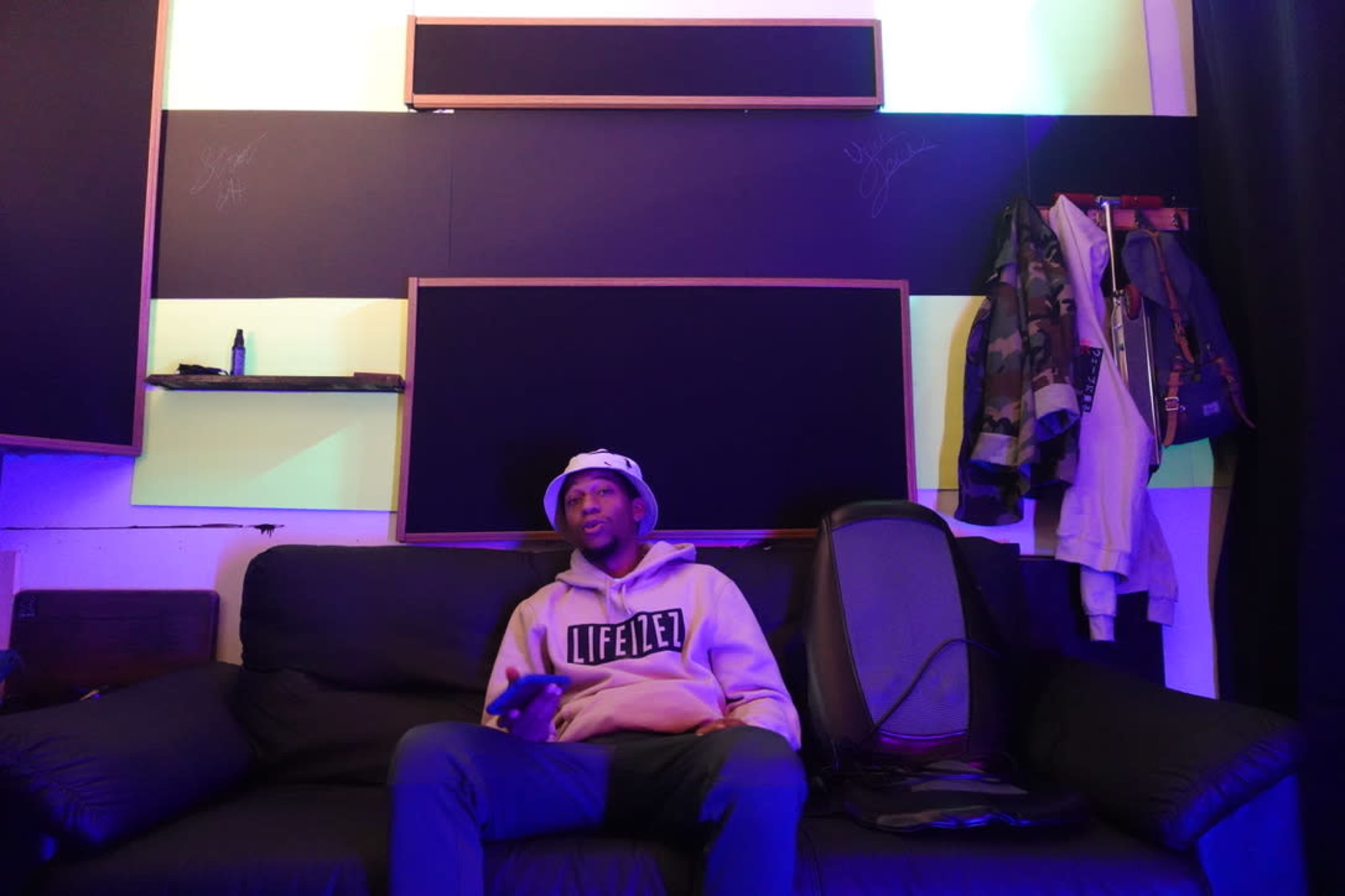 A person sits on a black sofa in a brightly lit room with green and blue walls, surrounded by clothing and audio equipment.