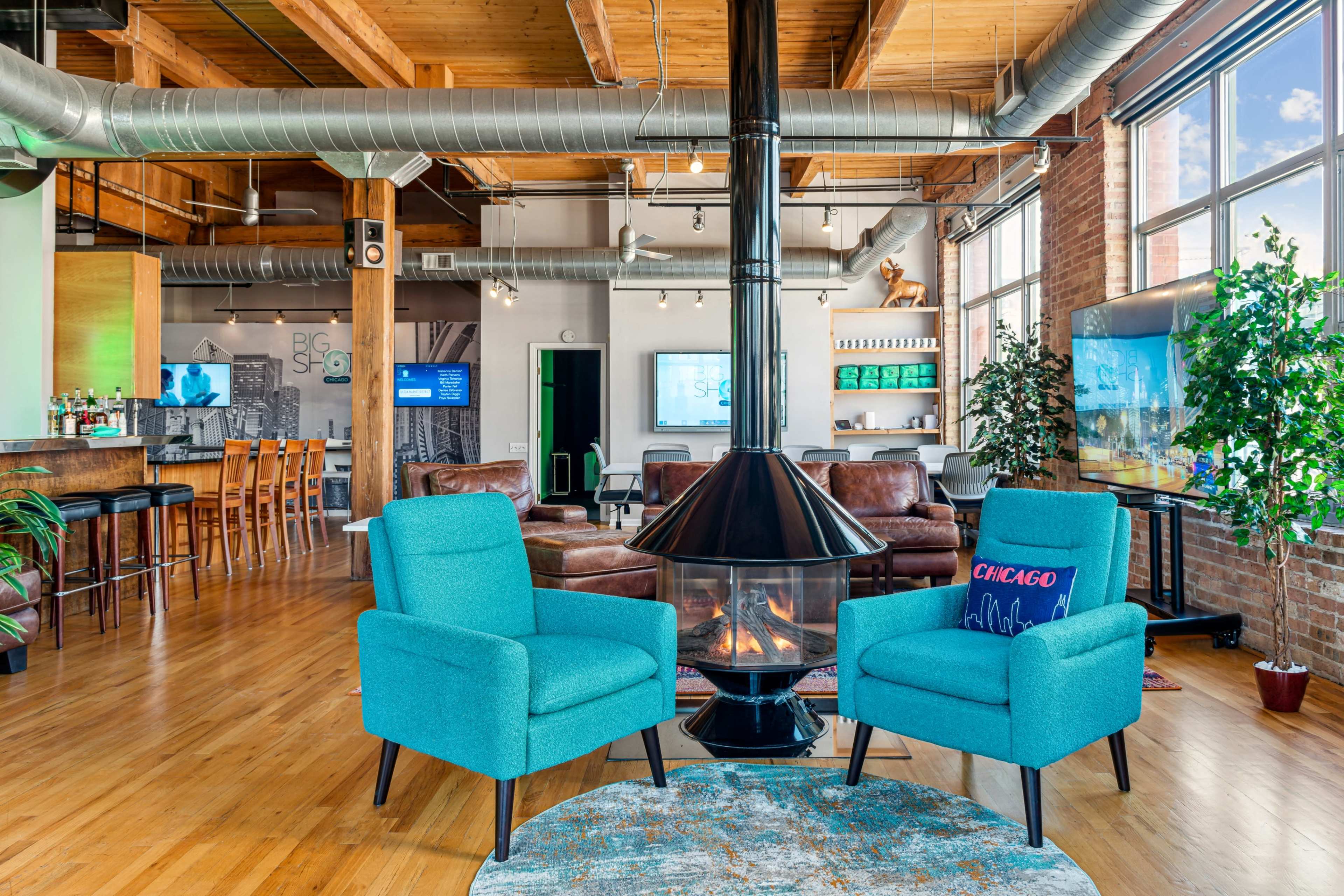 A modern loft-style lounge area featuring two turquoise armchairs, a circular rug, a central fireplace, and a bar with wooden stools in a spacious, well-lit environment.