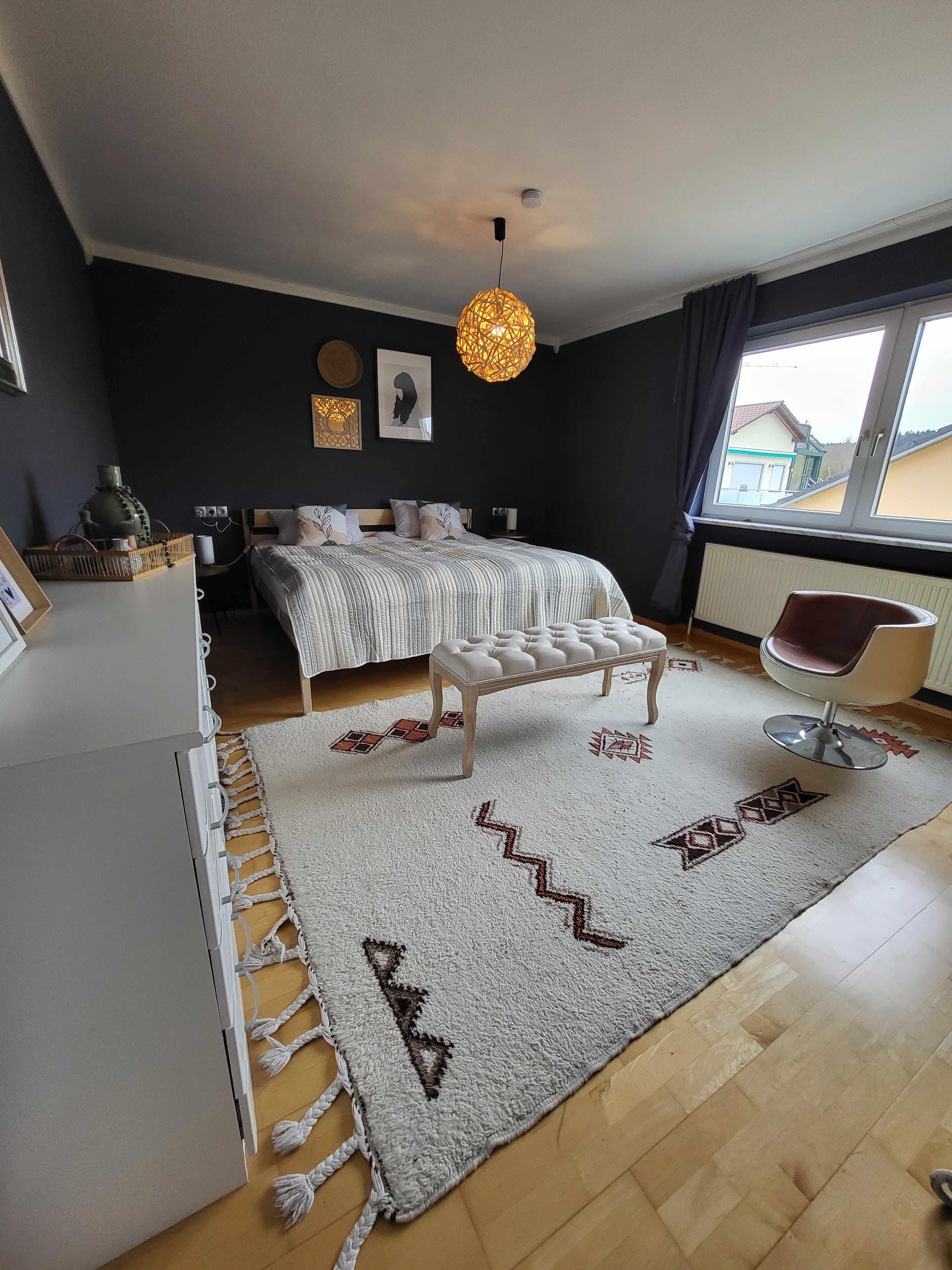 A modern bedroom featuring a striped bed, a decorative bench, a large area rug, and a sleek chair beside a window.