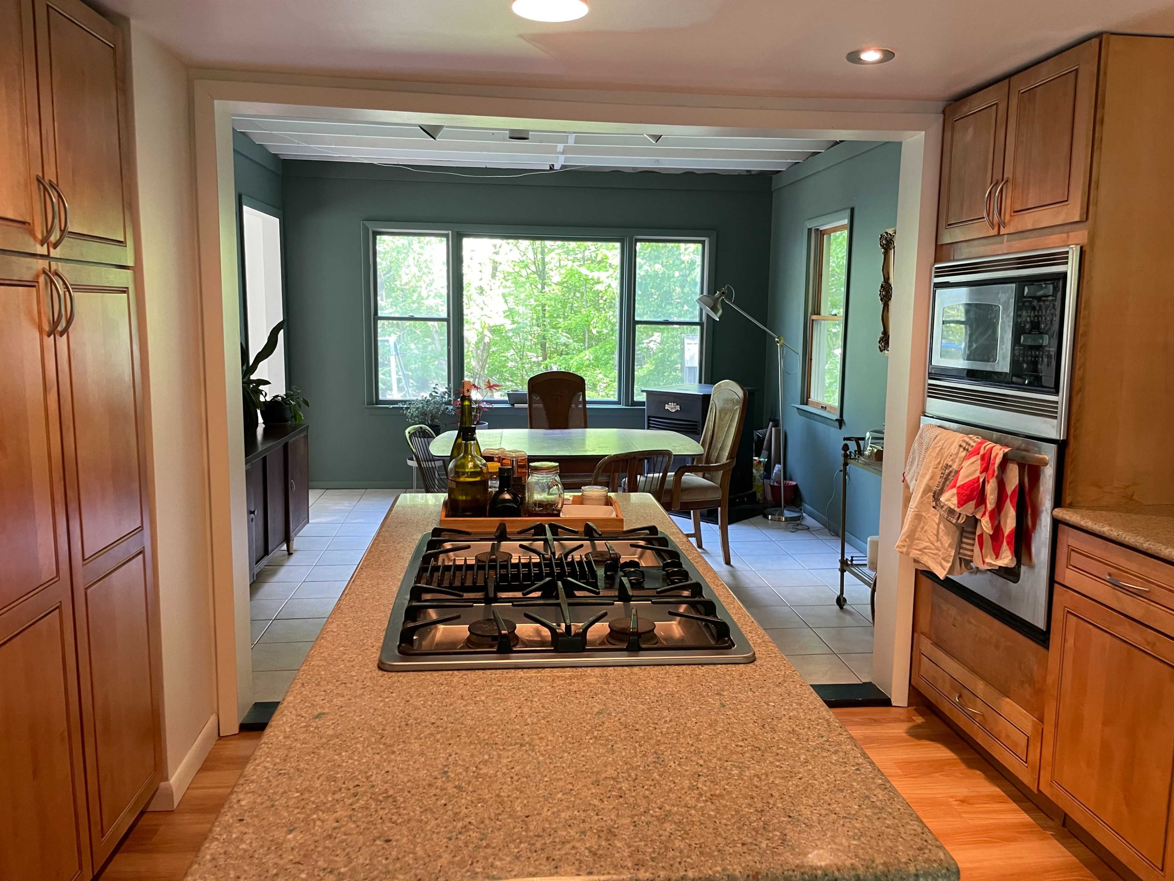 A kitchen with wooden cabinetry and a gas stove, leading to a dining area with large windows and a table set for meals.