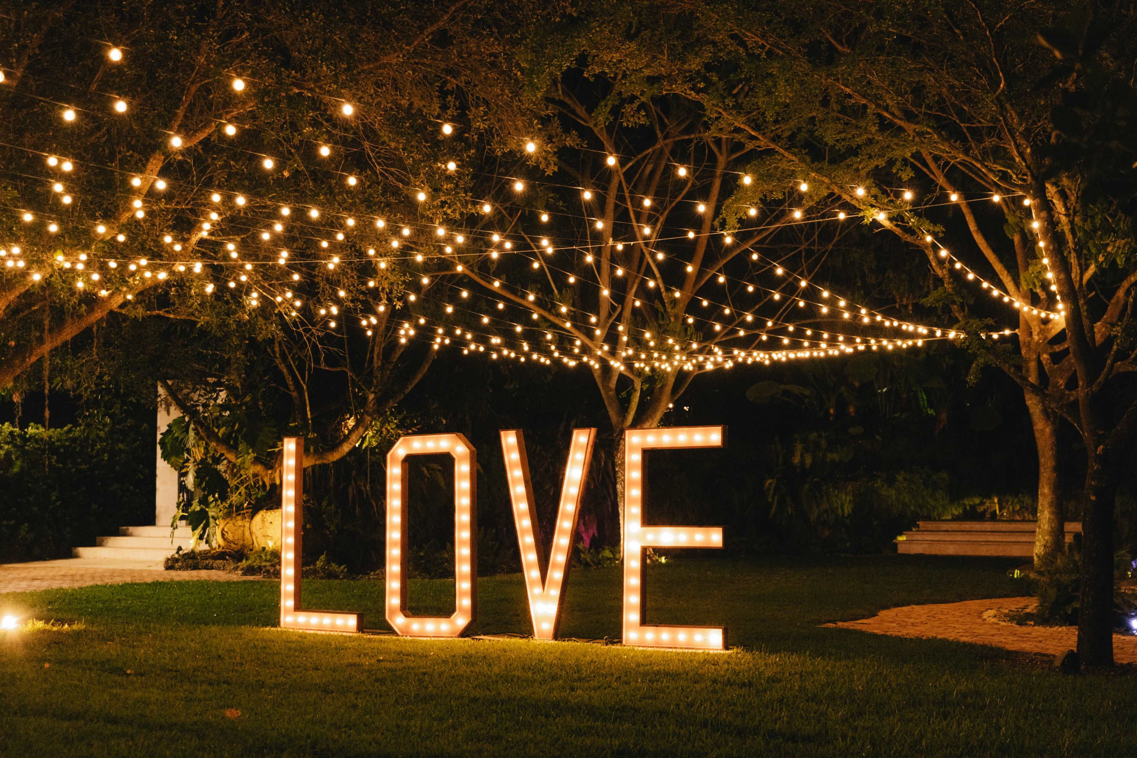 Large illuminated letters spelling "LOVE" are set in a garden adorned with string lights hanging from trees, creating a warm evening atmosphere.