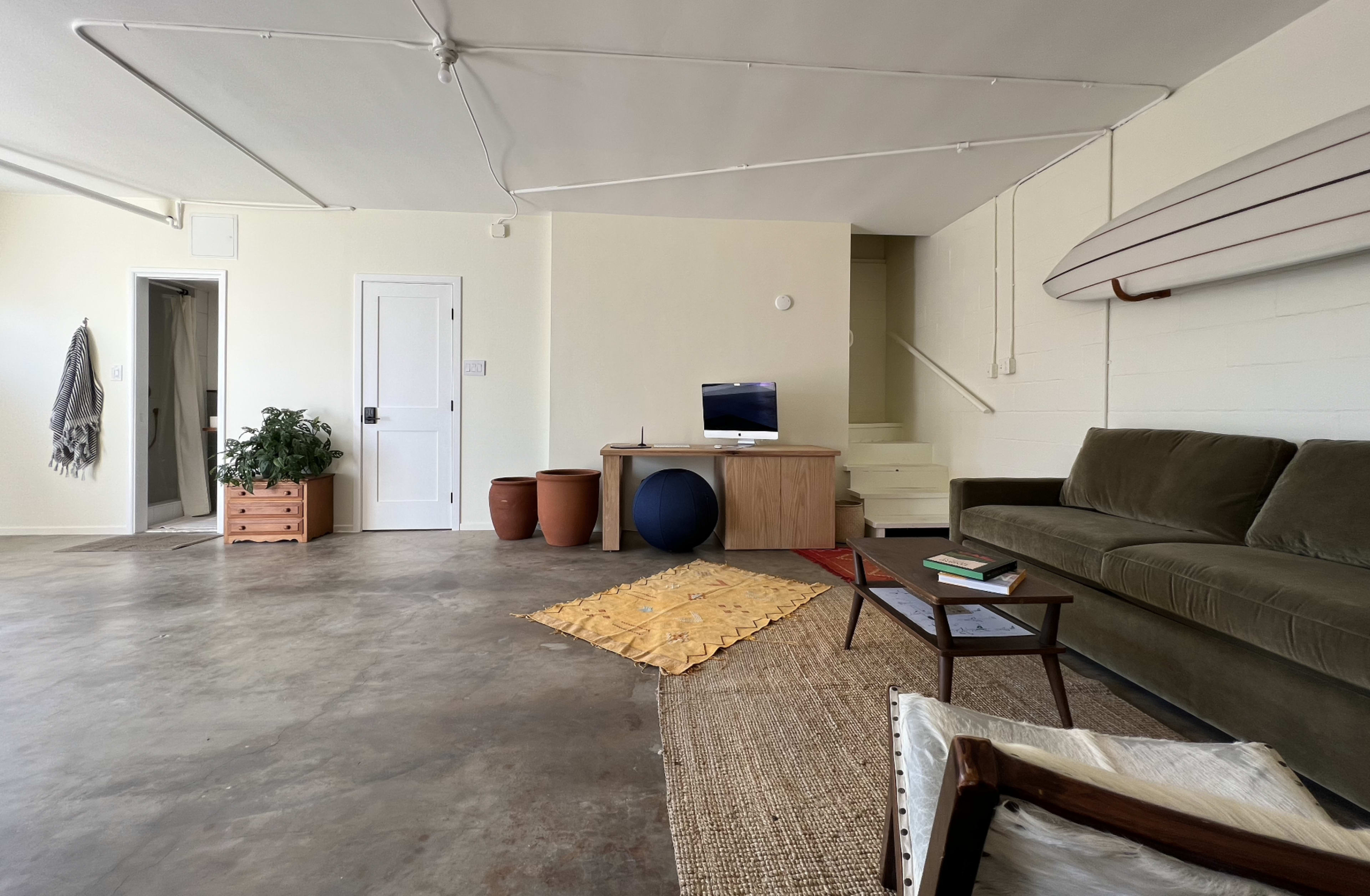 The image shows a minimalist living space with a green sofa, a wooden coffee table, decorative items, and a surfboard mounted on the wall.