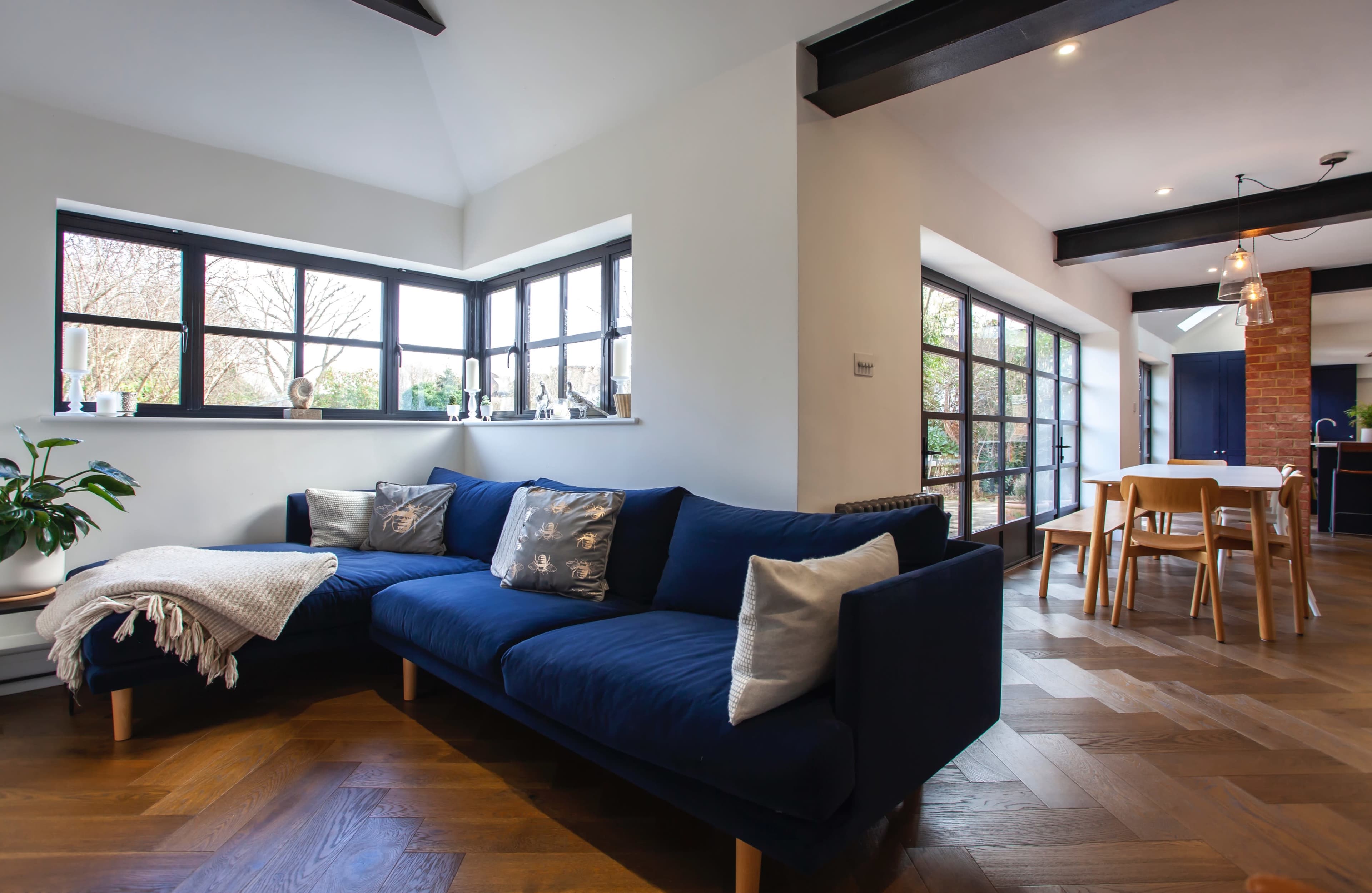 A modern living room features a navy blue sectional sofa, large windows allowing natural light, and a dining area with a wooden table.