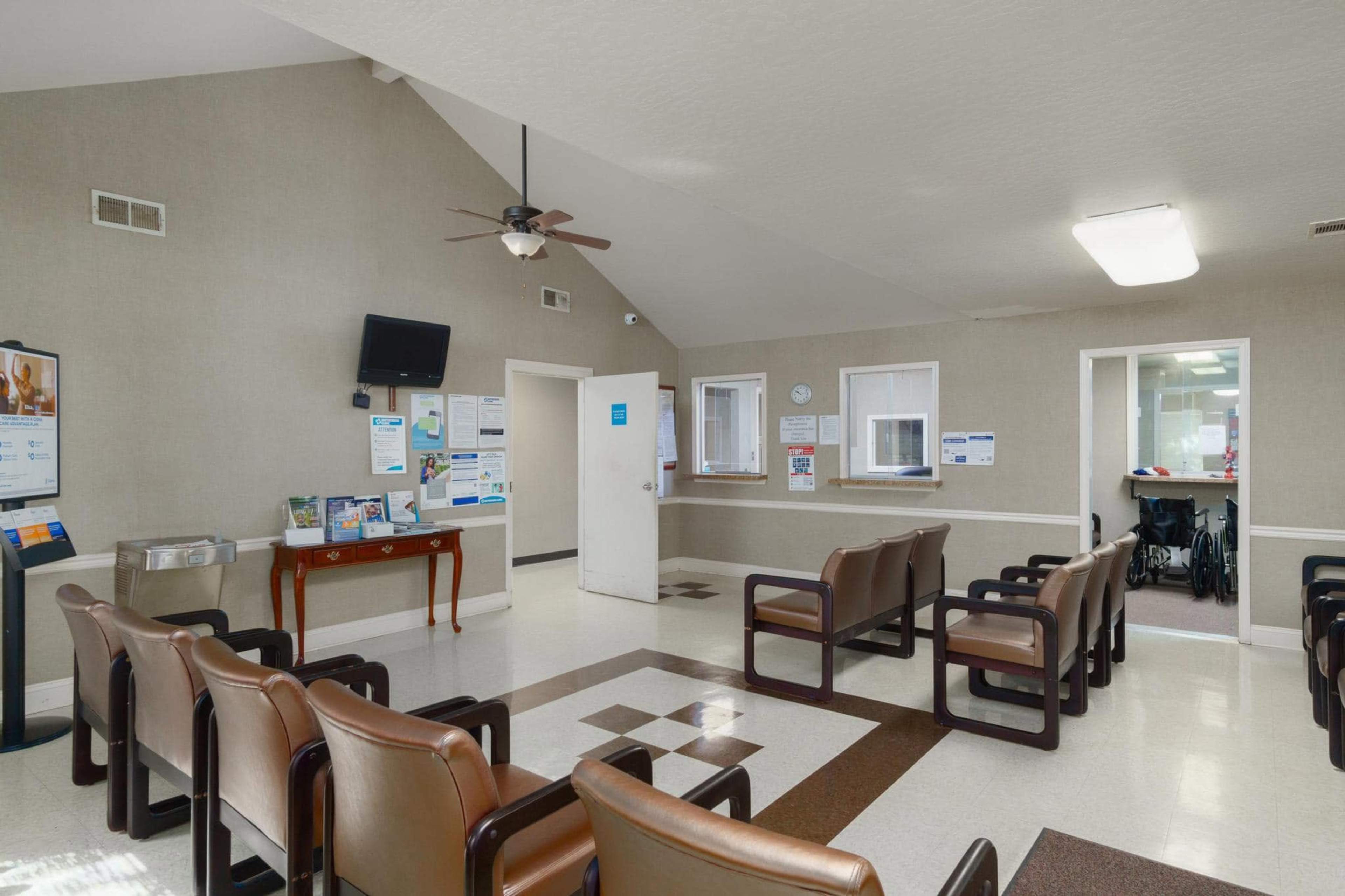 The image shows a waiting room with several rows of brown chairs, a table displaying informational brochures, and a television mounted on the wall.