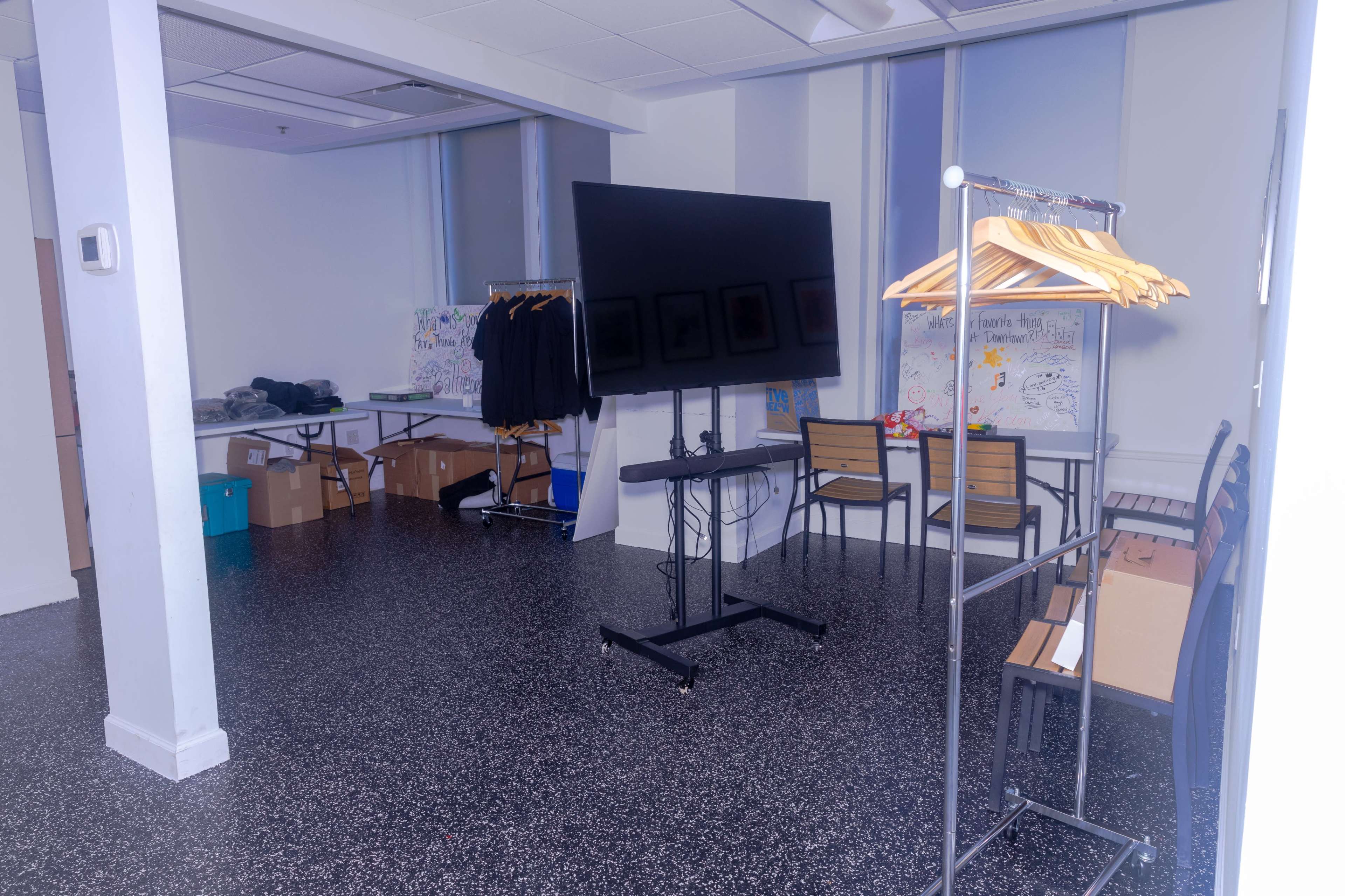 The image shows an empty room with a large screen on a stand, a clothing rack with hangers, chairs, and several boxes stacked along the walls.