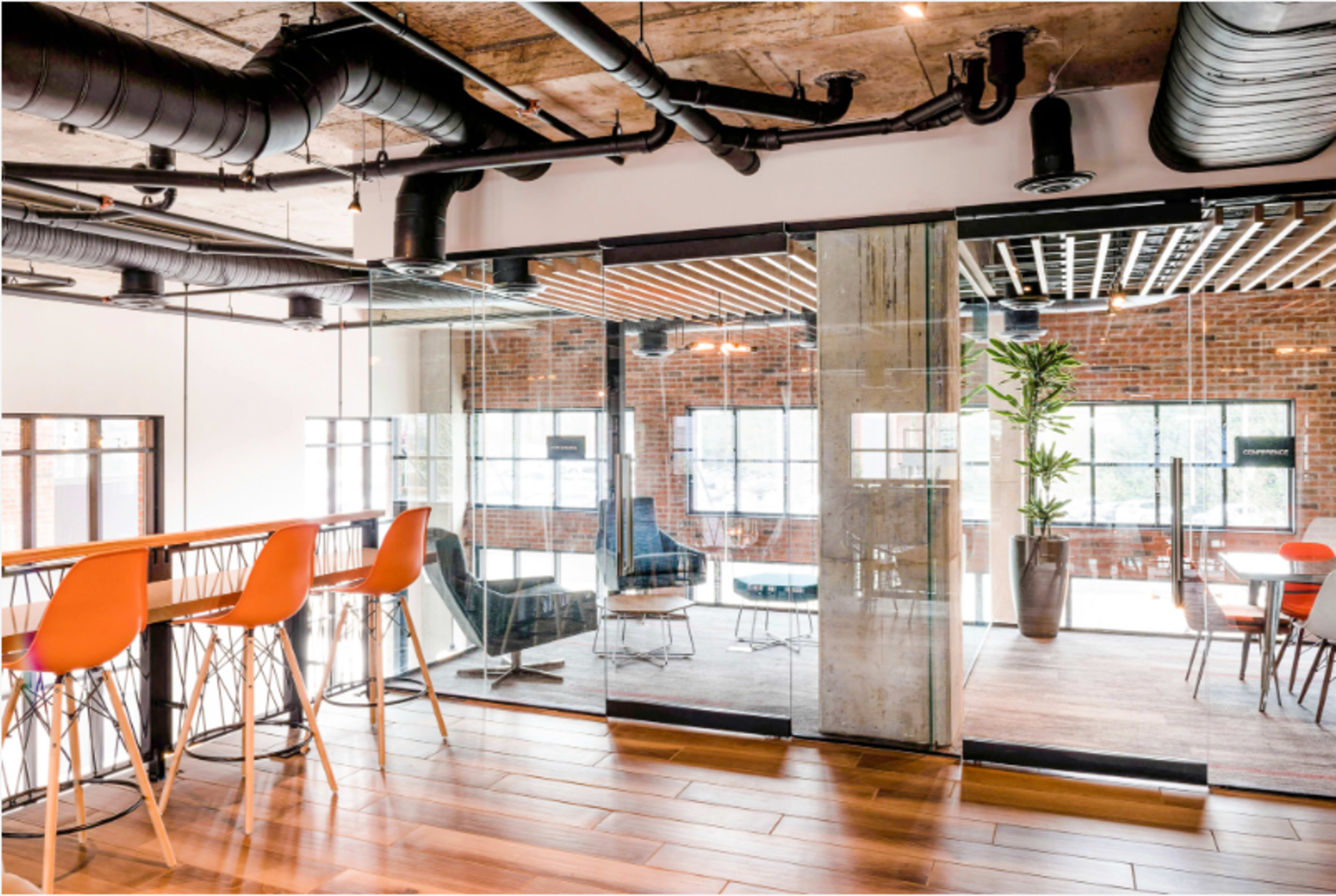 The image shows a modern office space with a mix of glass walls, wooden flooring, and colorful seating arrangements.