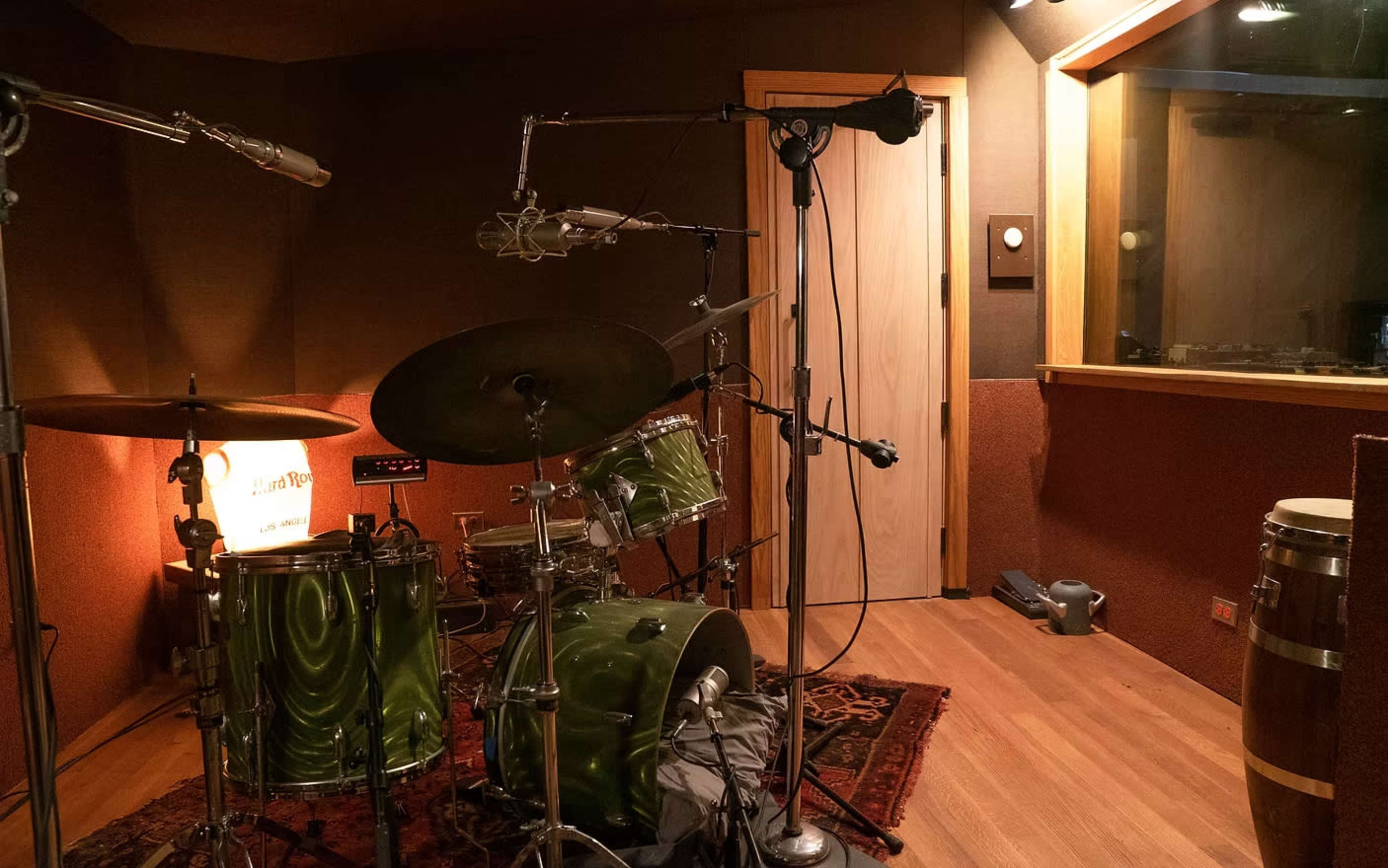 The image shows a recording studio with a green drum set, microphones arranged, and a glass-walled control room in the background.