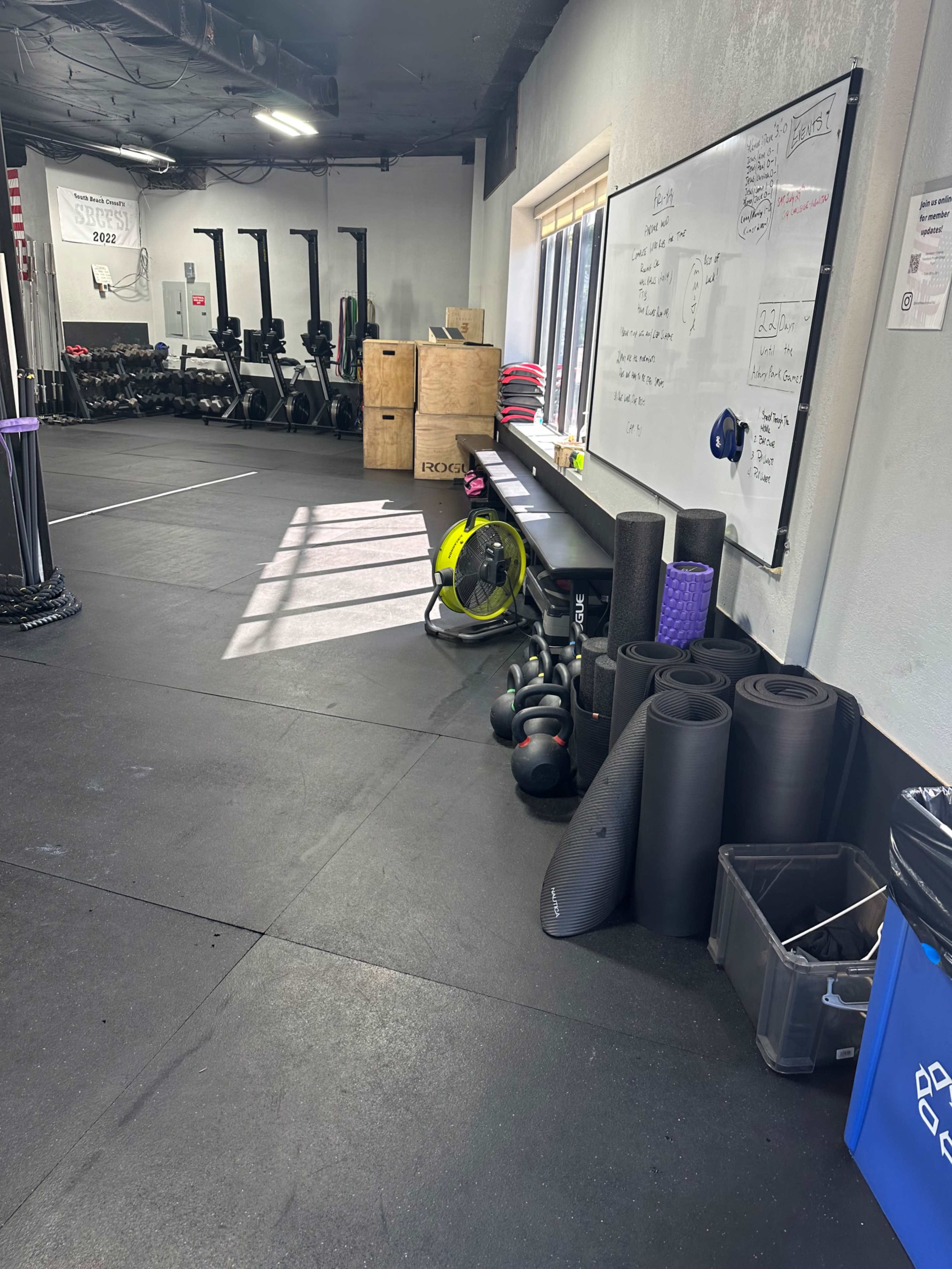 The image shows a fitness area with gym equipment, including kettlebells, mats, and exercise bikes, along with a whiteboard on the wall.