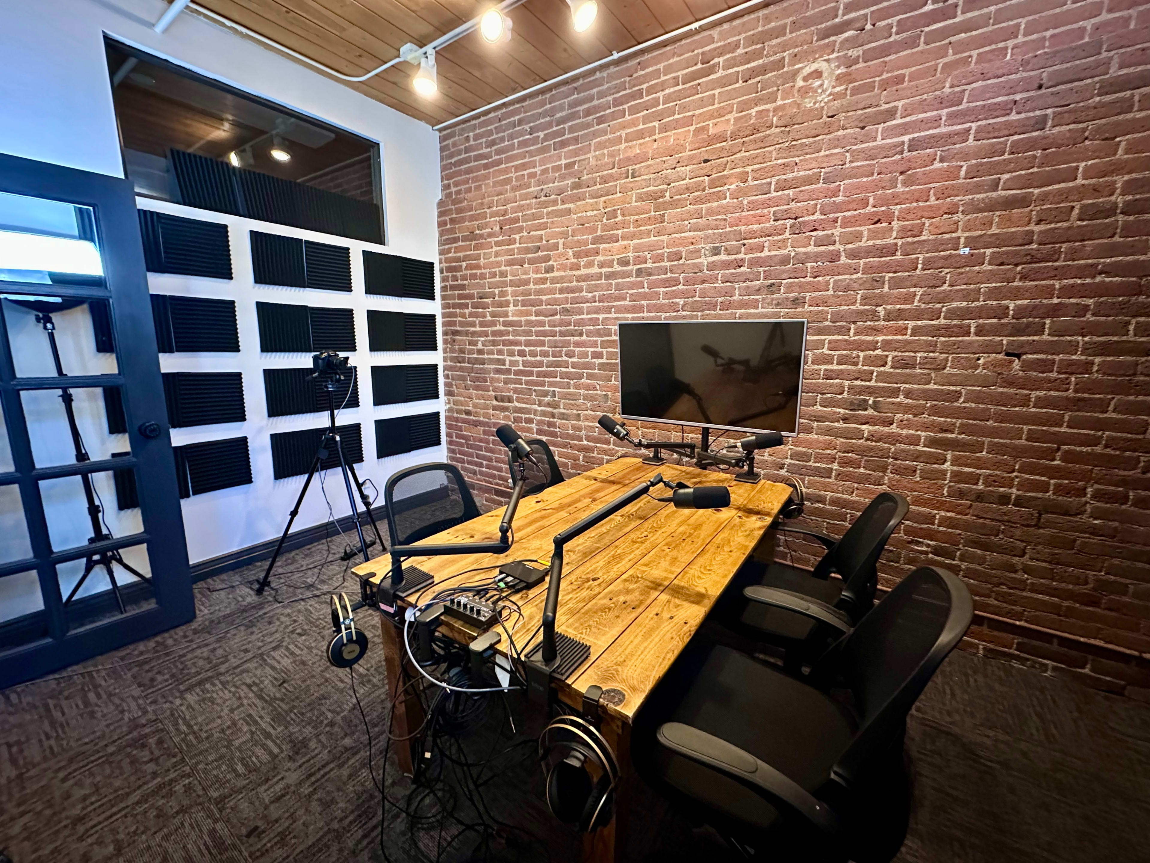 The image shows a recording studio with a wooden table, several microphones, and a monitor against a brick wall, along with soundproofing panels and a camera setup in the background.