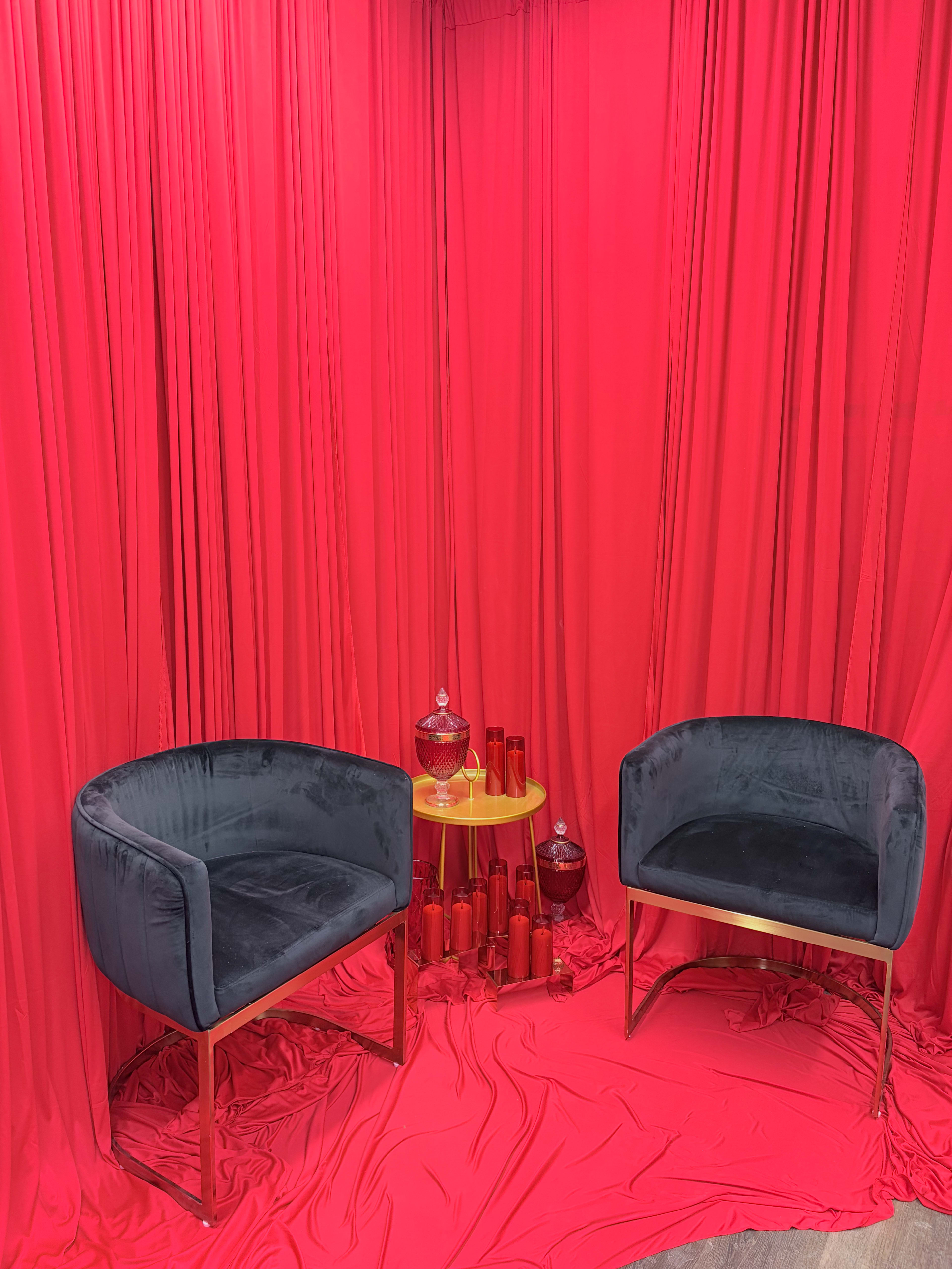 Two dark upholstered chairs are positioned on a red fabric-covered floor, surrounded by flowing red curtains and decorative glass objects on a gold table.