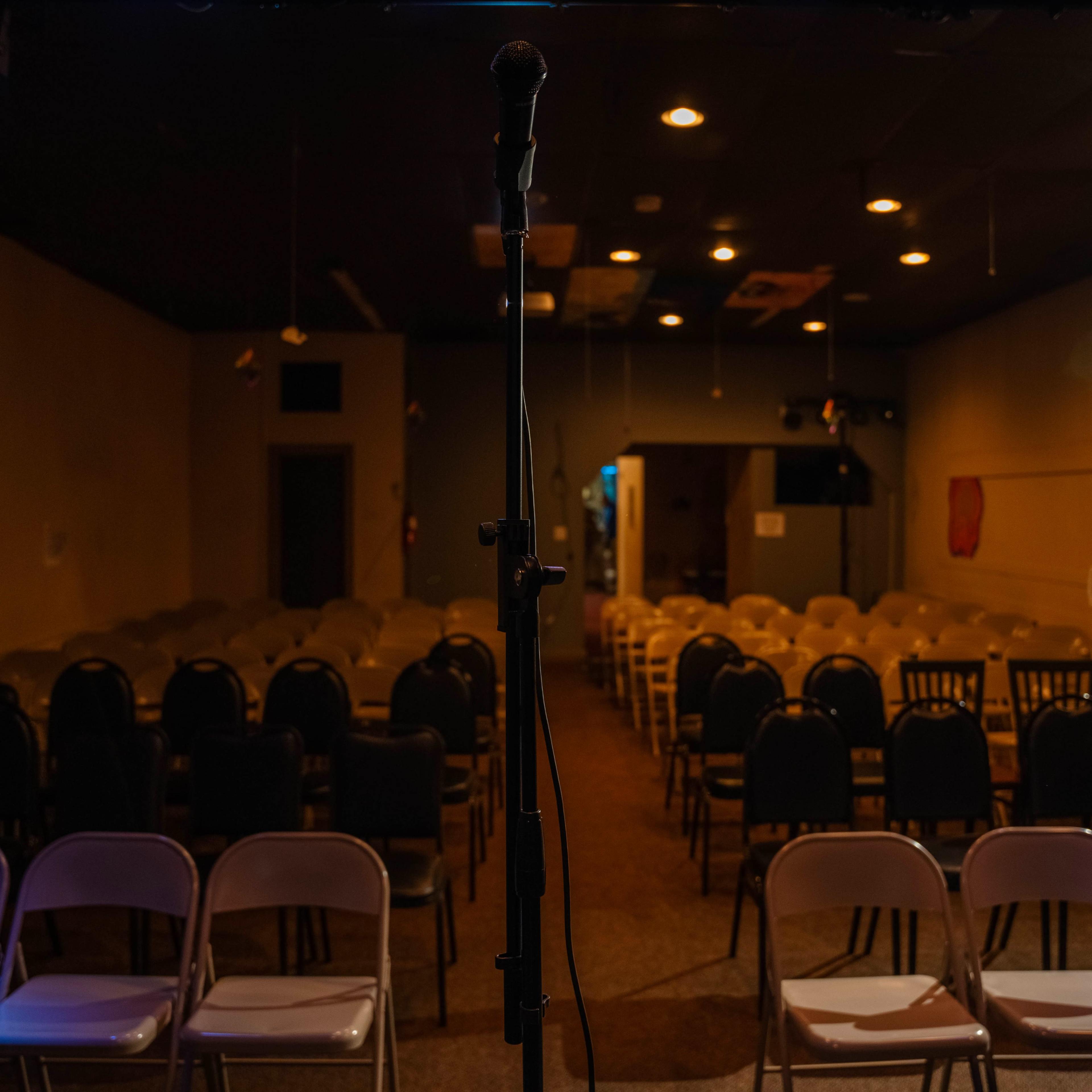 A microphone stands at the front of a dimly lit room filled with rows of empty chairs set up for an event.