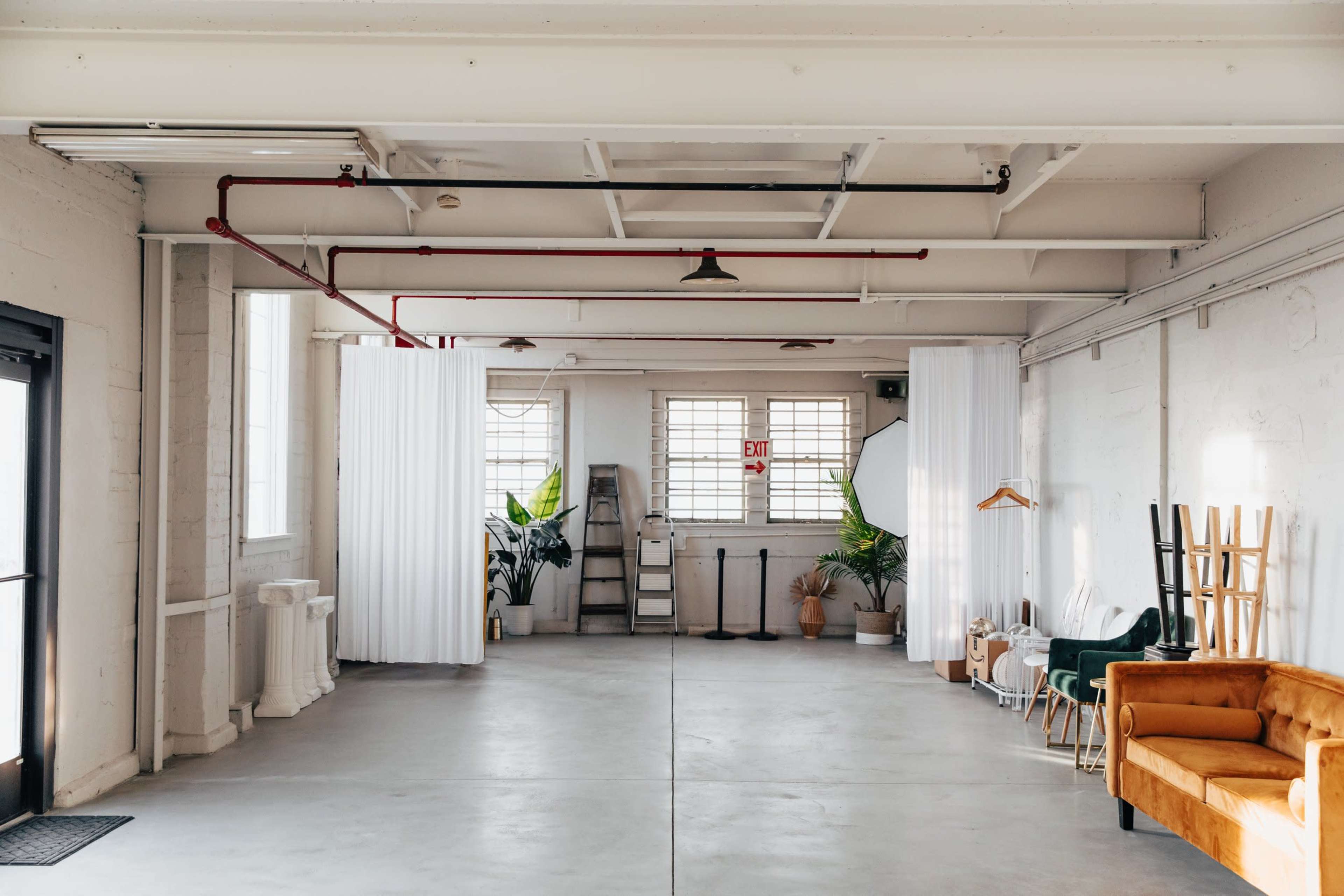 A spacious, minimalistic room with white walls, concrete flooring, and large windows, featuring plants, light curtains, and seating arrangements along the sides.