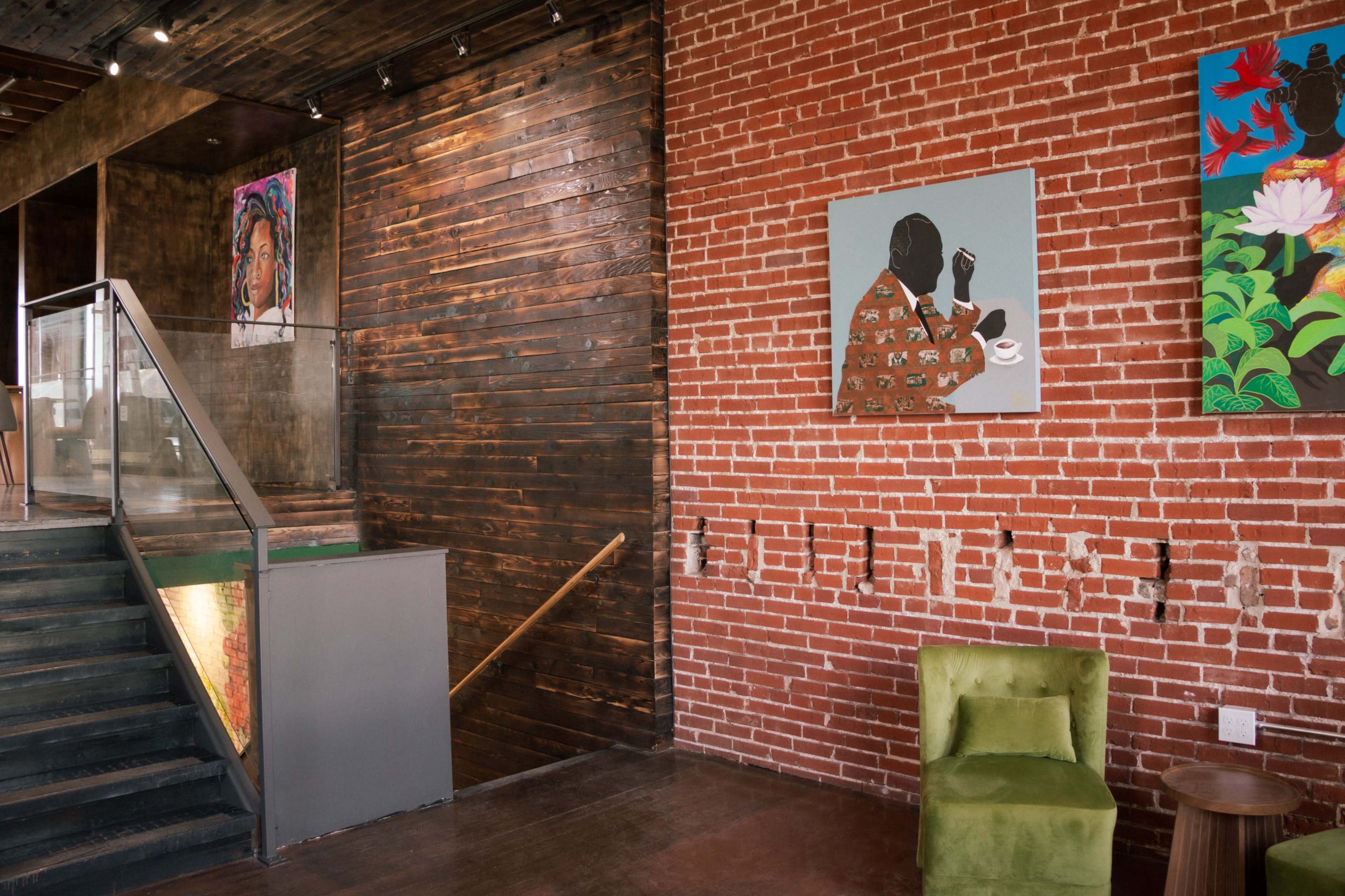 The image shows an interior scene featuring a staircase against a brick wall, adorned with colorful artwork, and a green chair in the corner.