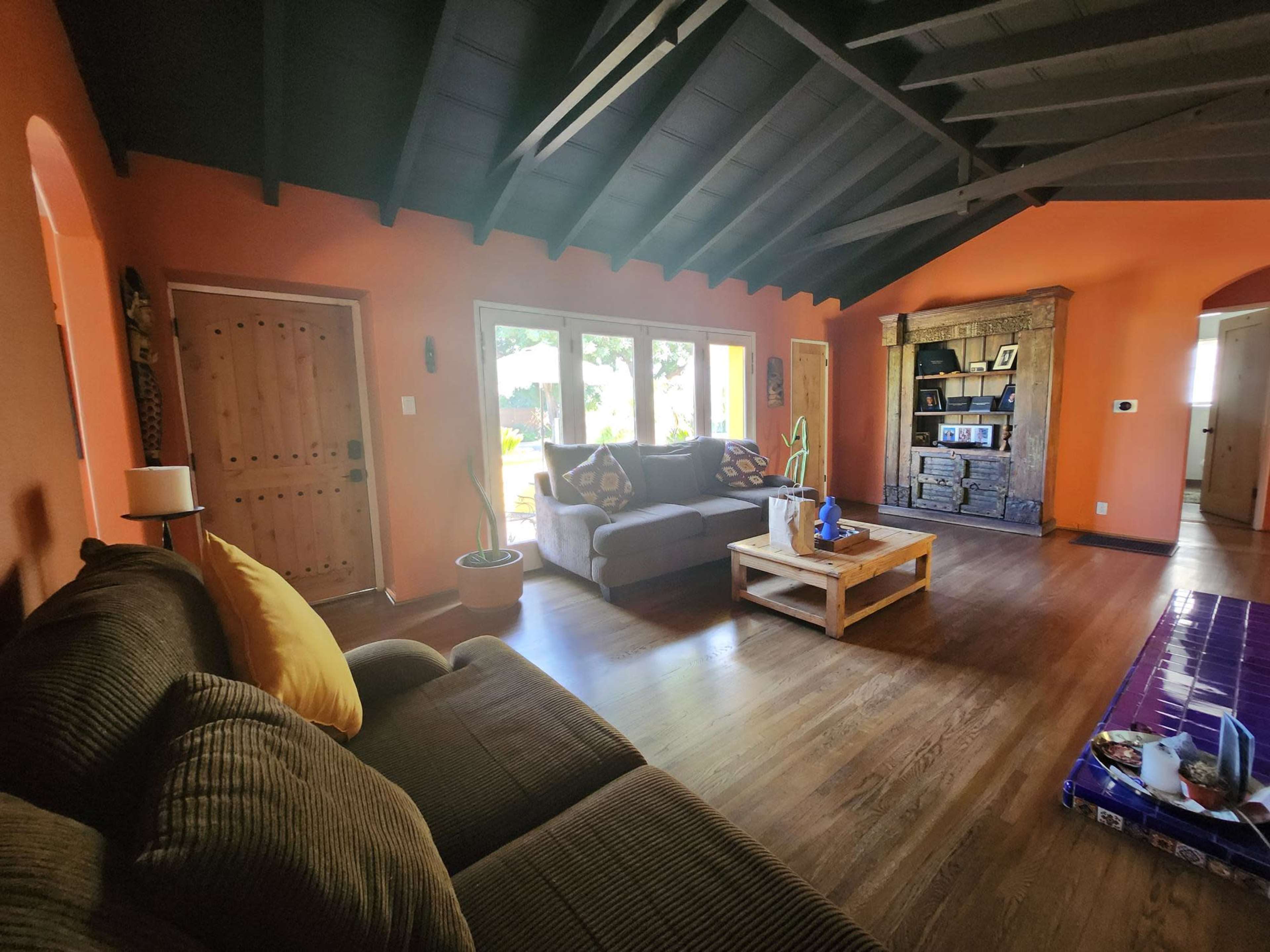 The image shows a living room with a brown couch, a wooden coffee table, and large windows overlooking a garden, all under a sloped ceiling.