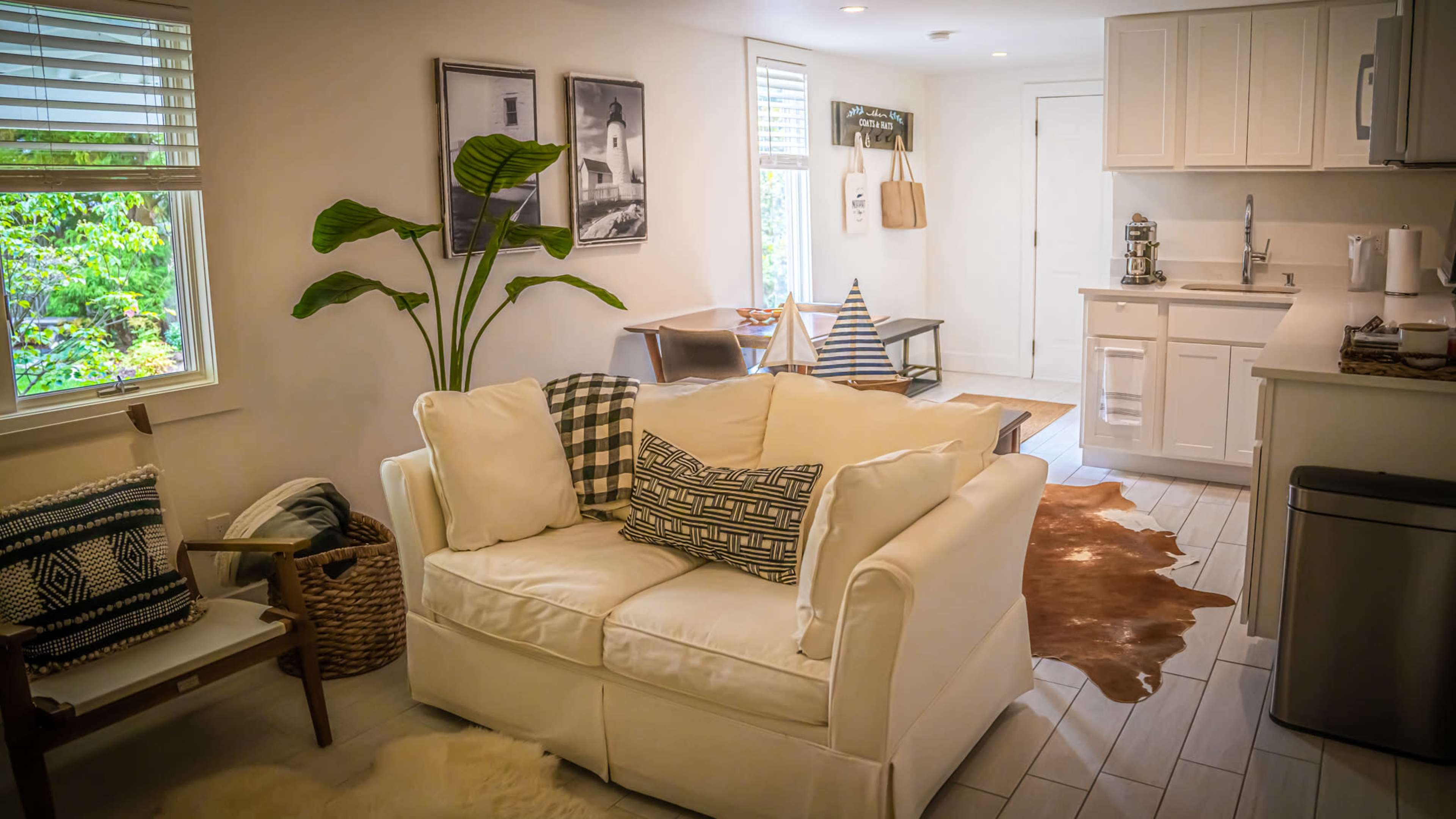 A bright, modern living area featuring a white couch with patterned pillows, a small dining area, and a kitchen in the background, all surrounded by greenery outside.