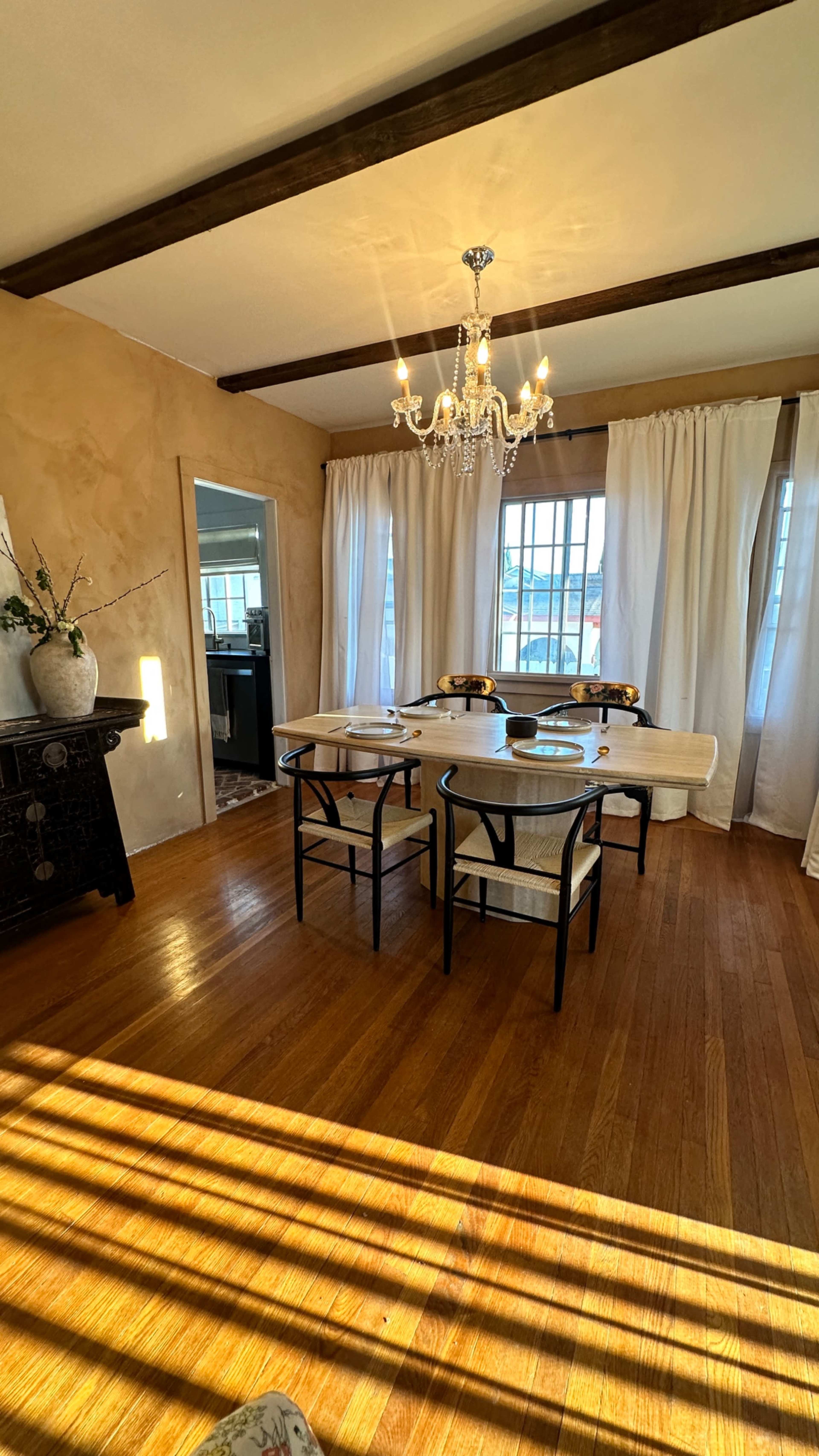 The dining area features a round table with chairs surrounded by large windows, soft curtains, and a chandelier overhead.