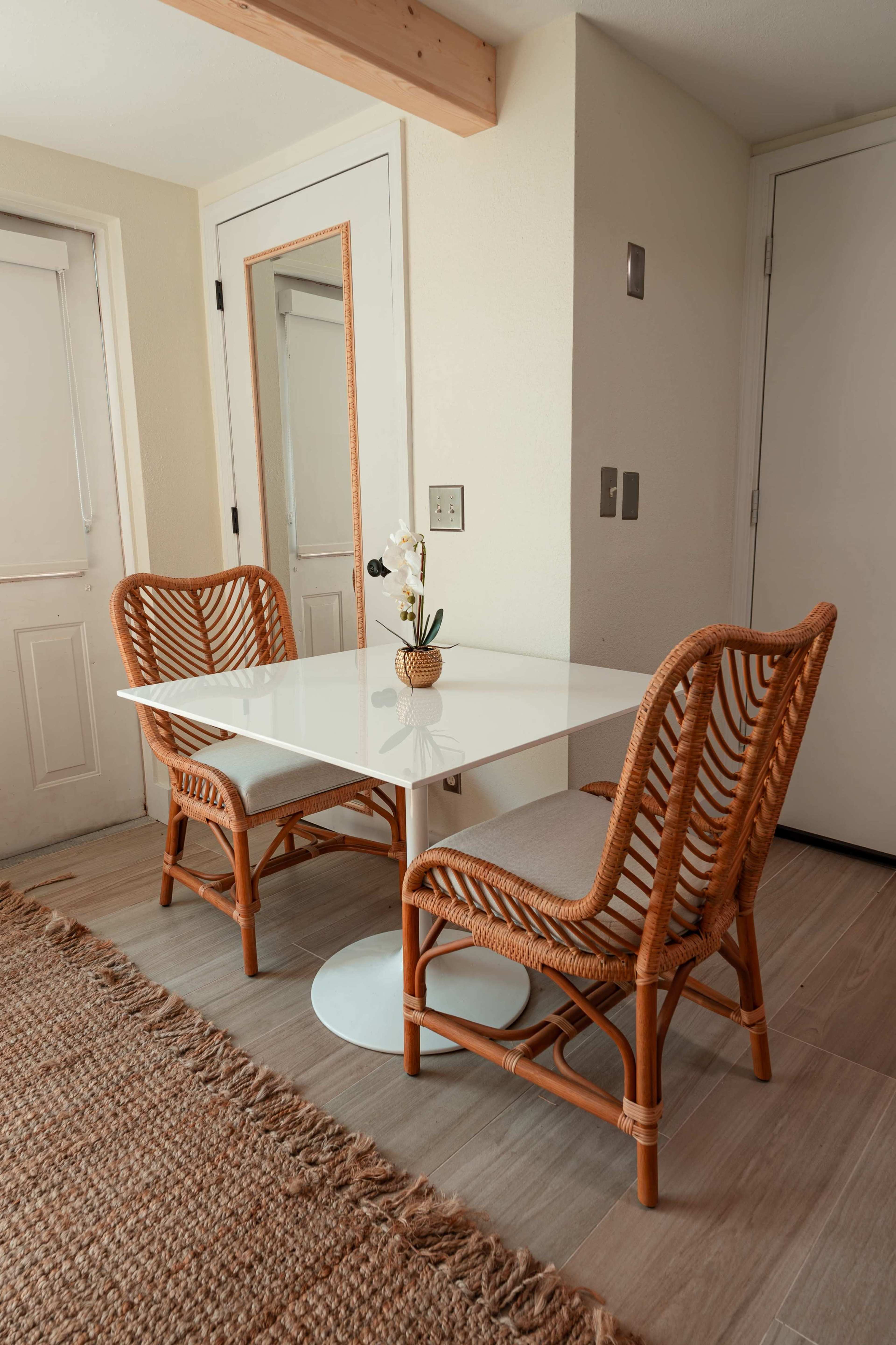 A small dining area features a white table with two rattan chairs and a potted plant, accompanied by a mirror on the wall.