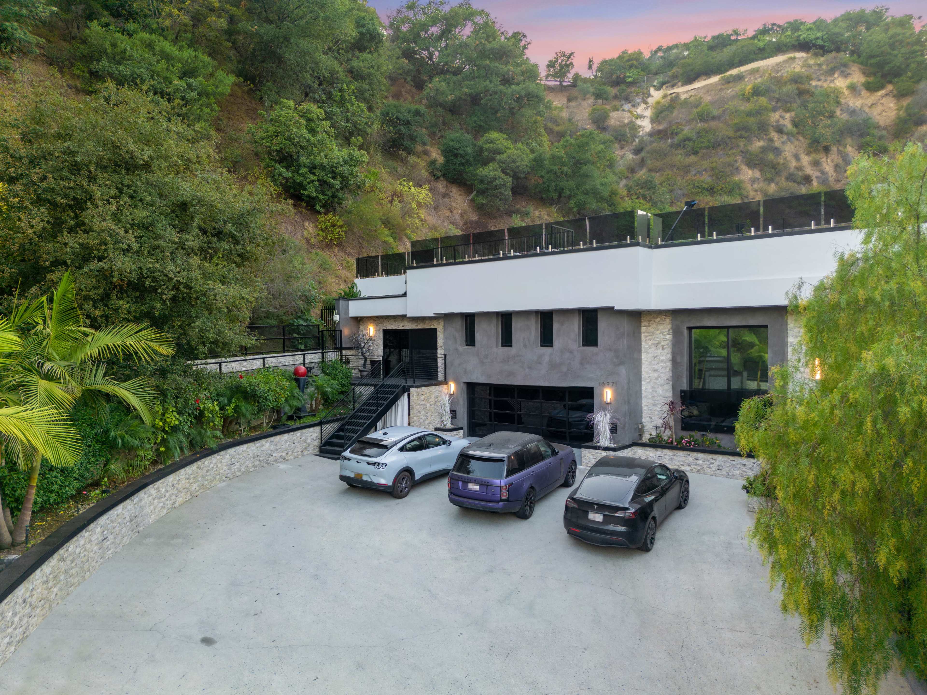 A modern multi-story house is surrounded by greenery and features a spacious parking area with four cars.