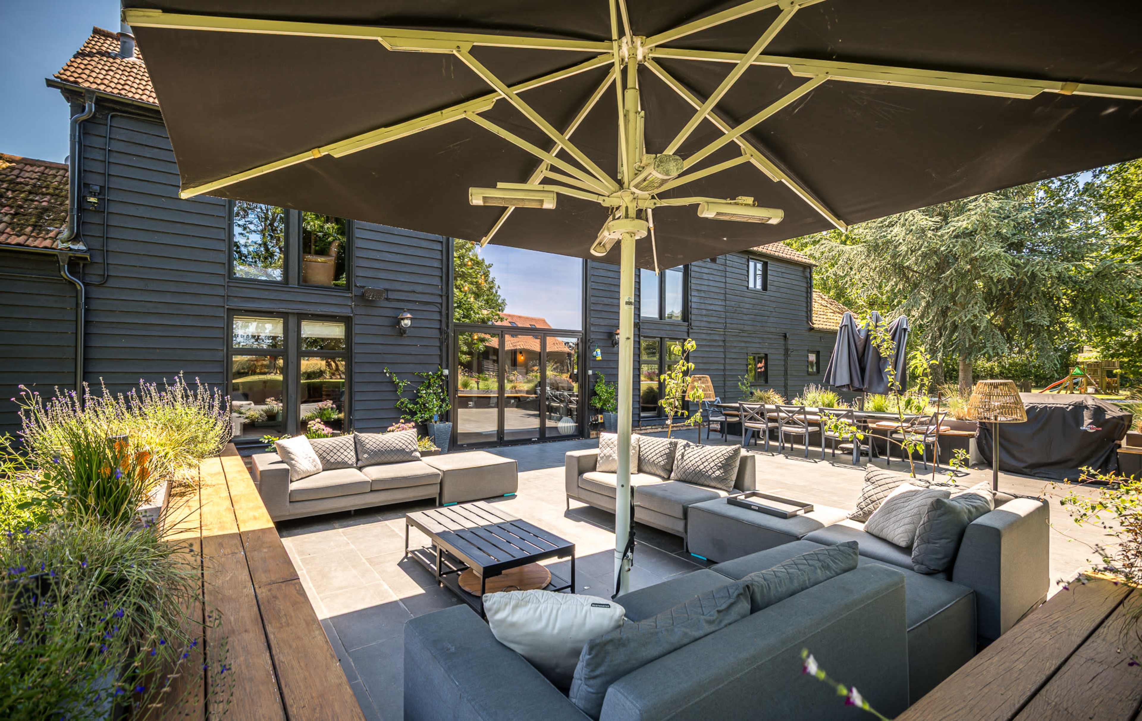 A spacious outdoor patio features modern seating arrangements, an umbrella, and views of a contemporary house surrounded by greenery.