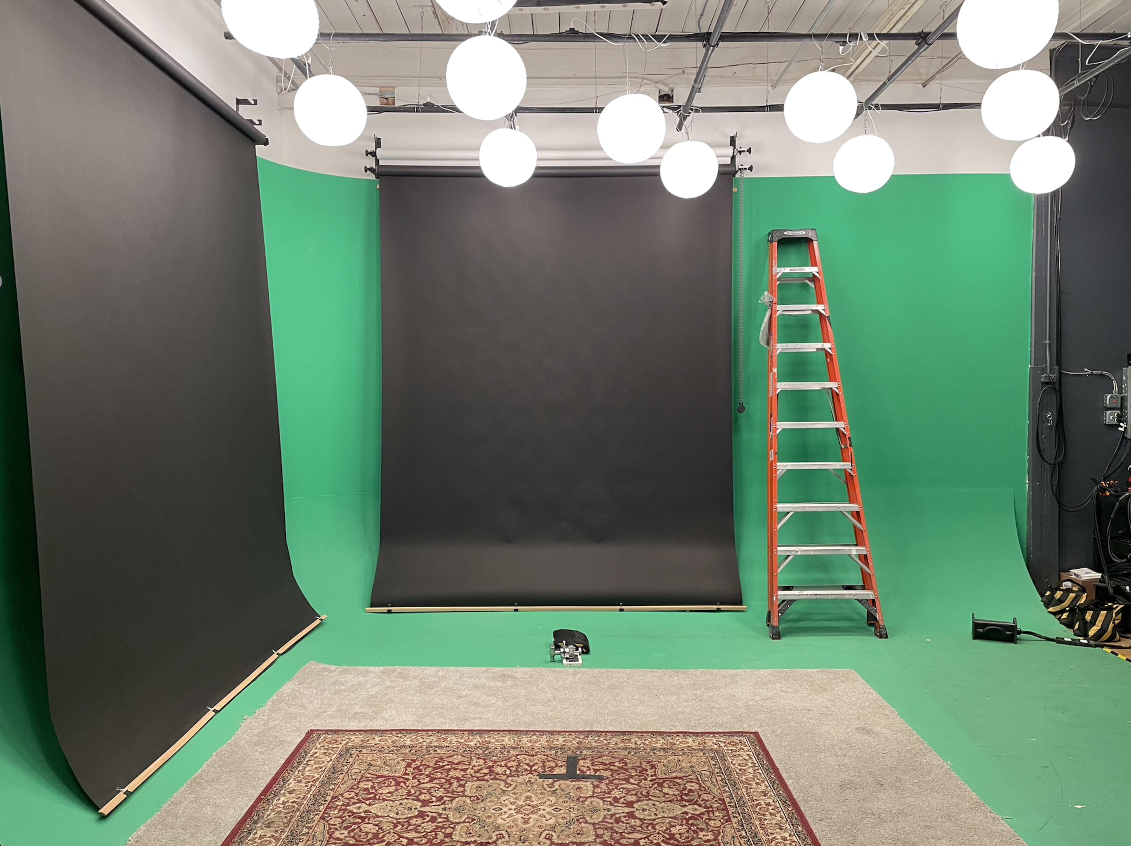 The scene features a green screen studio with two black backdrops, a ladder, and hanging lights above a carpeted area.