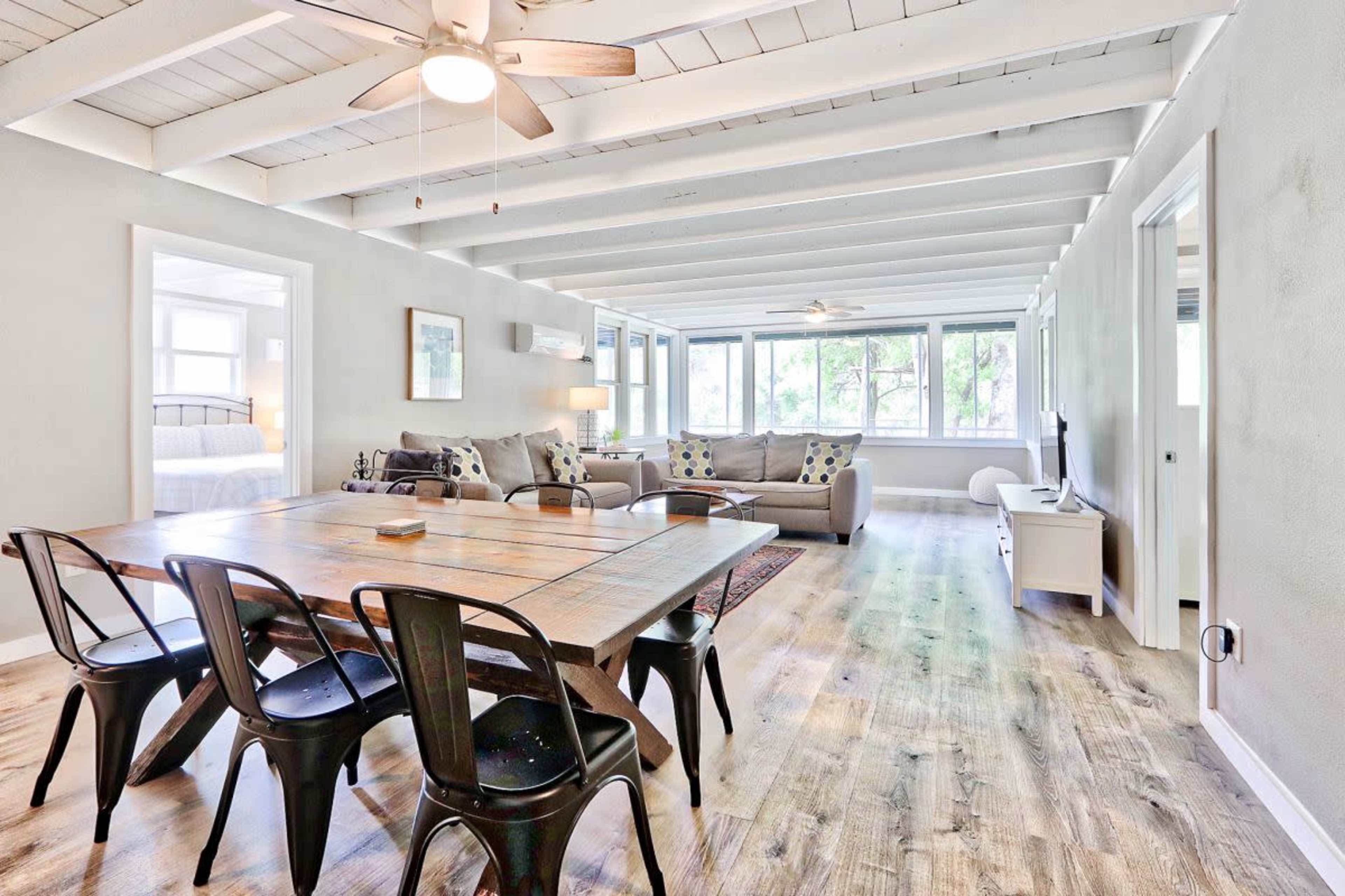 A spacious living area features a dining table with black chairs, a sofa, and large windows providing ample natural light.