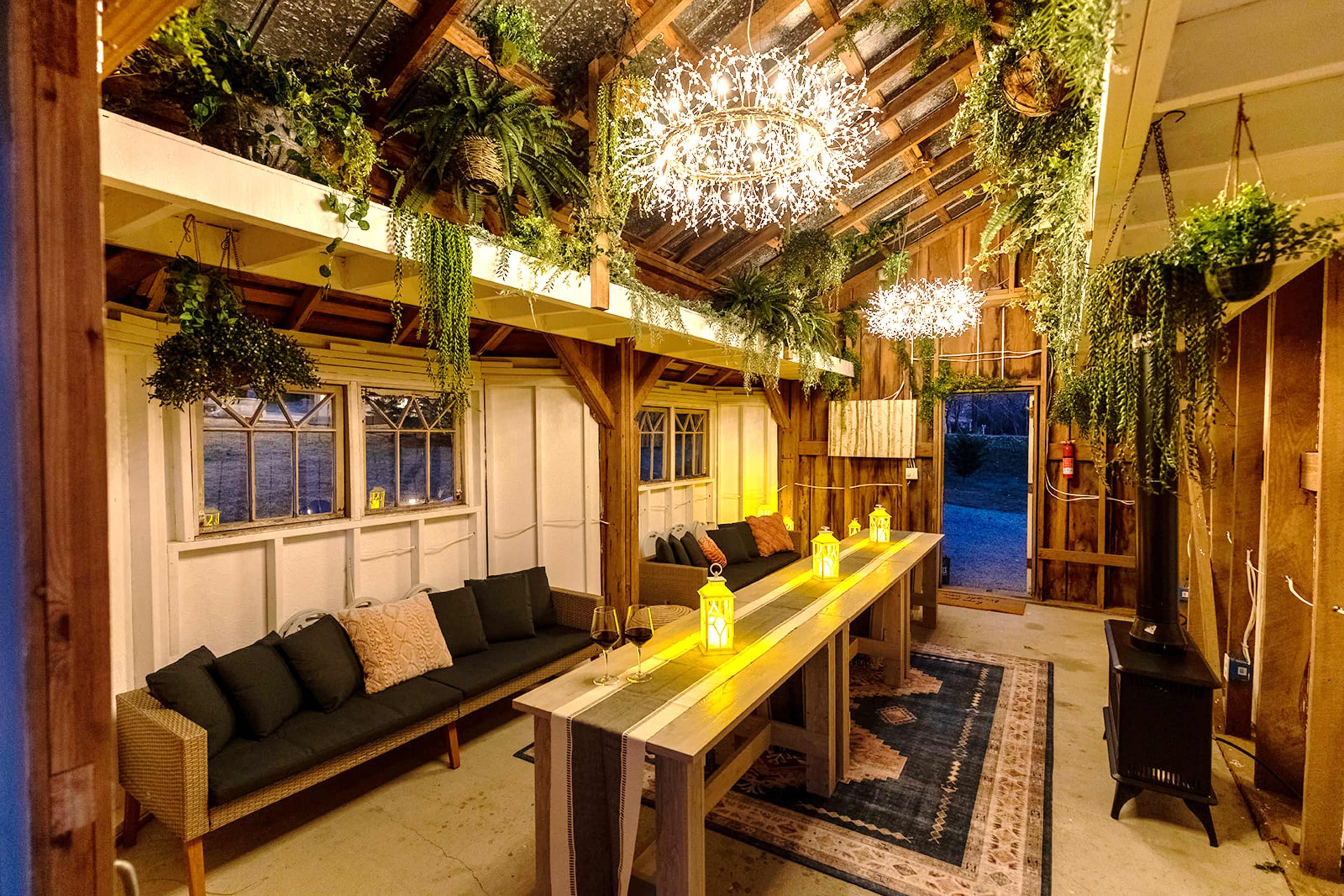 A rustic indoor space featuring a long wooden table, two stylish seating areas, and hanging lights amidst an array of plants.
