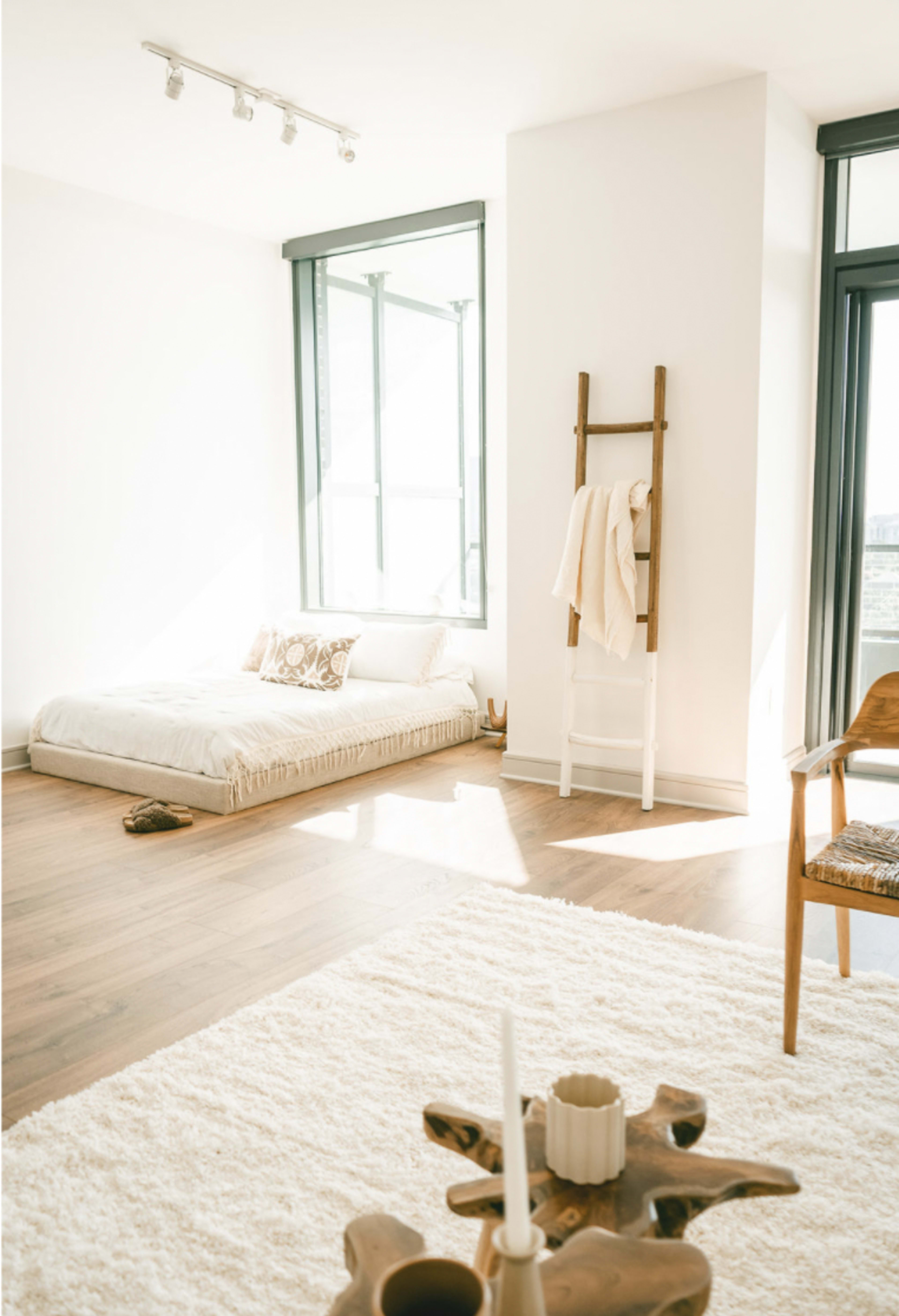A minimalist room features a low mattress on the floor, a wooden ladder holding towels, and a cozy, textured rug.