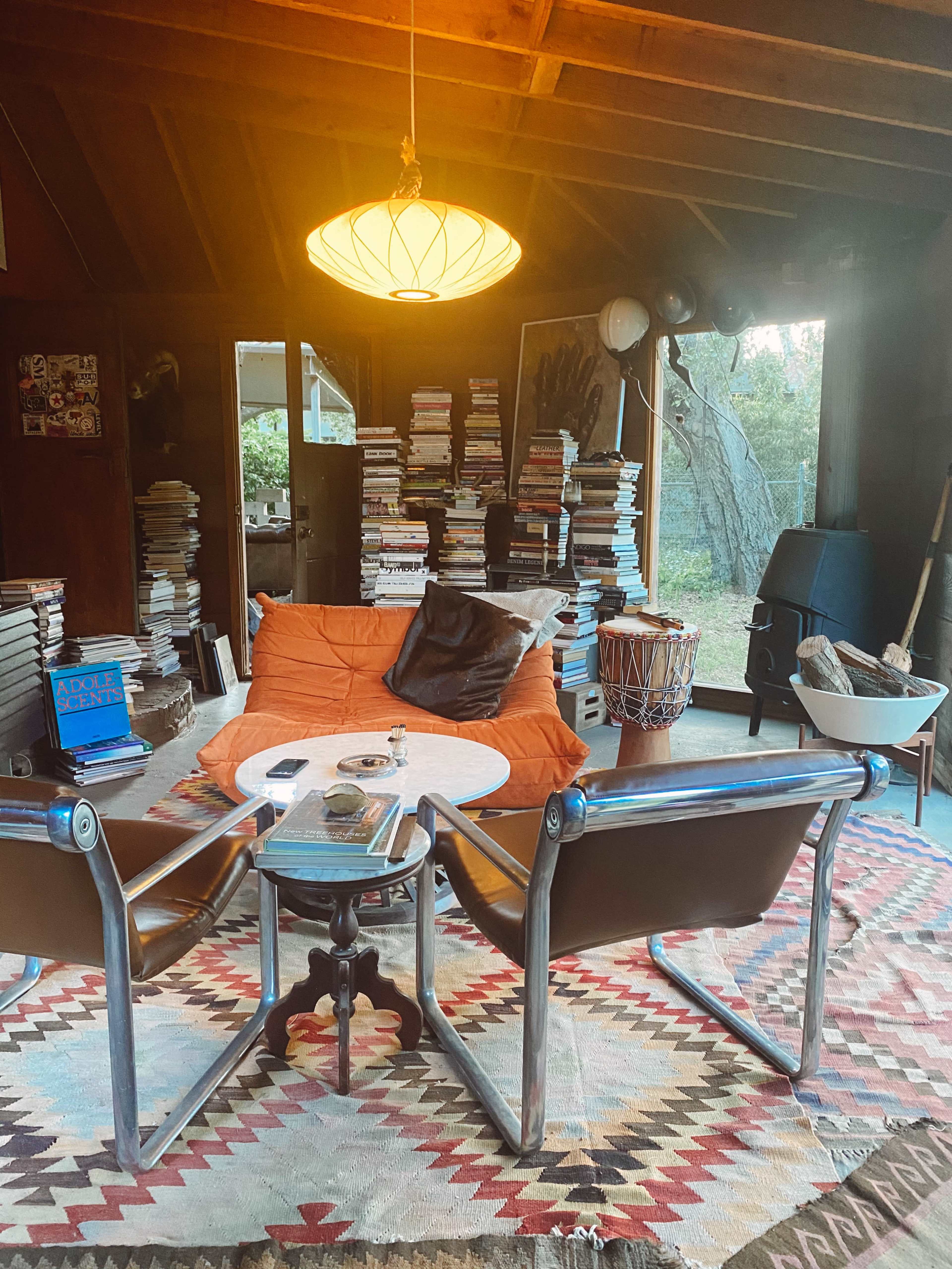 A cozy living room features a low table surrounded by chairs and a sofa, with stacks of books in the background and a large window letting in natural light.