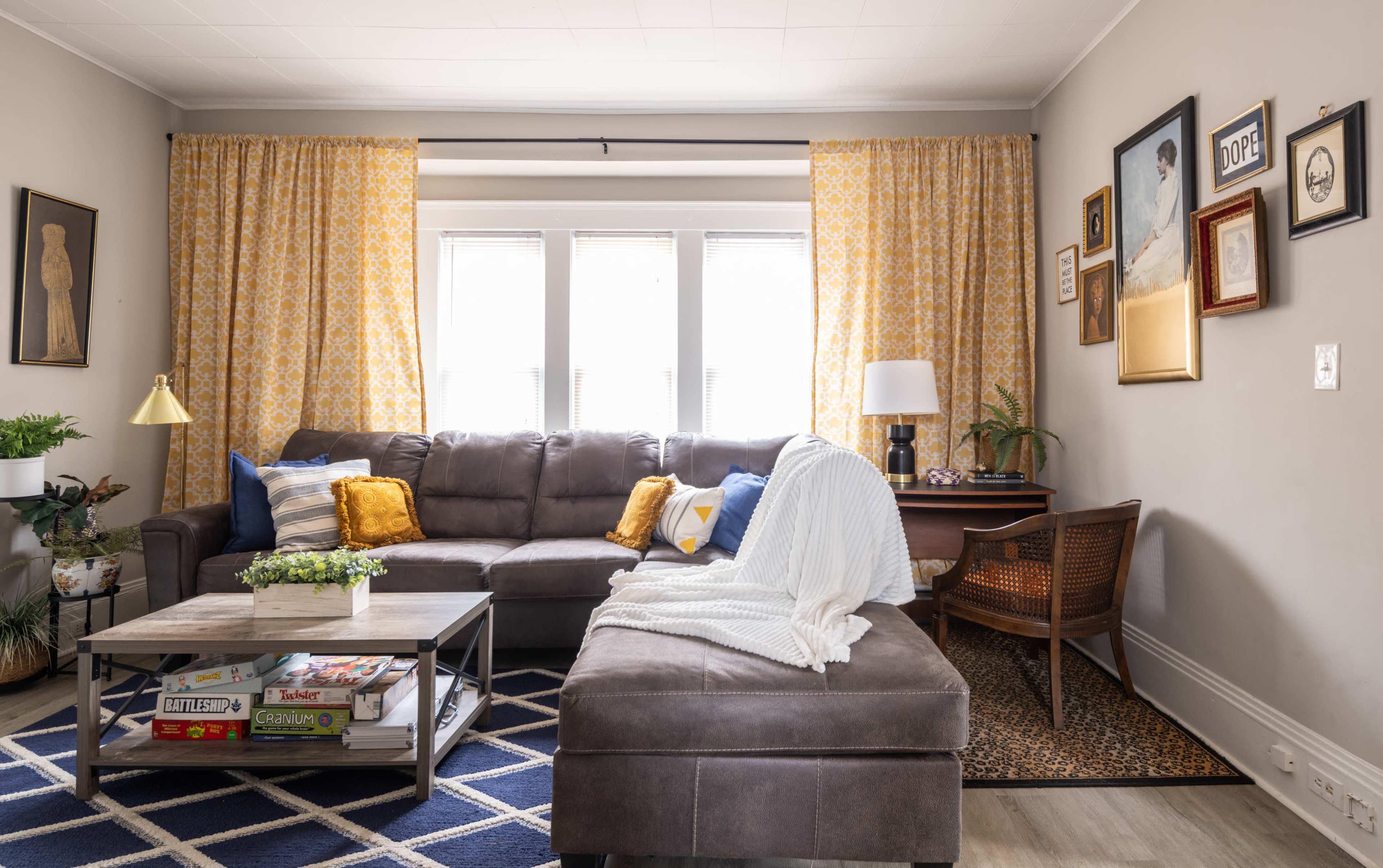 The living room features a sectional sofa with various cushions, a coffee table stacked with books, and large windows adorned with yellow patterned curtains.