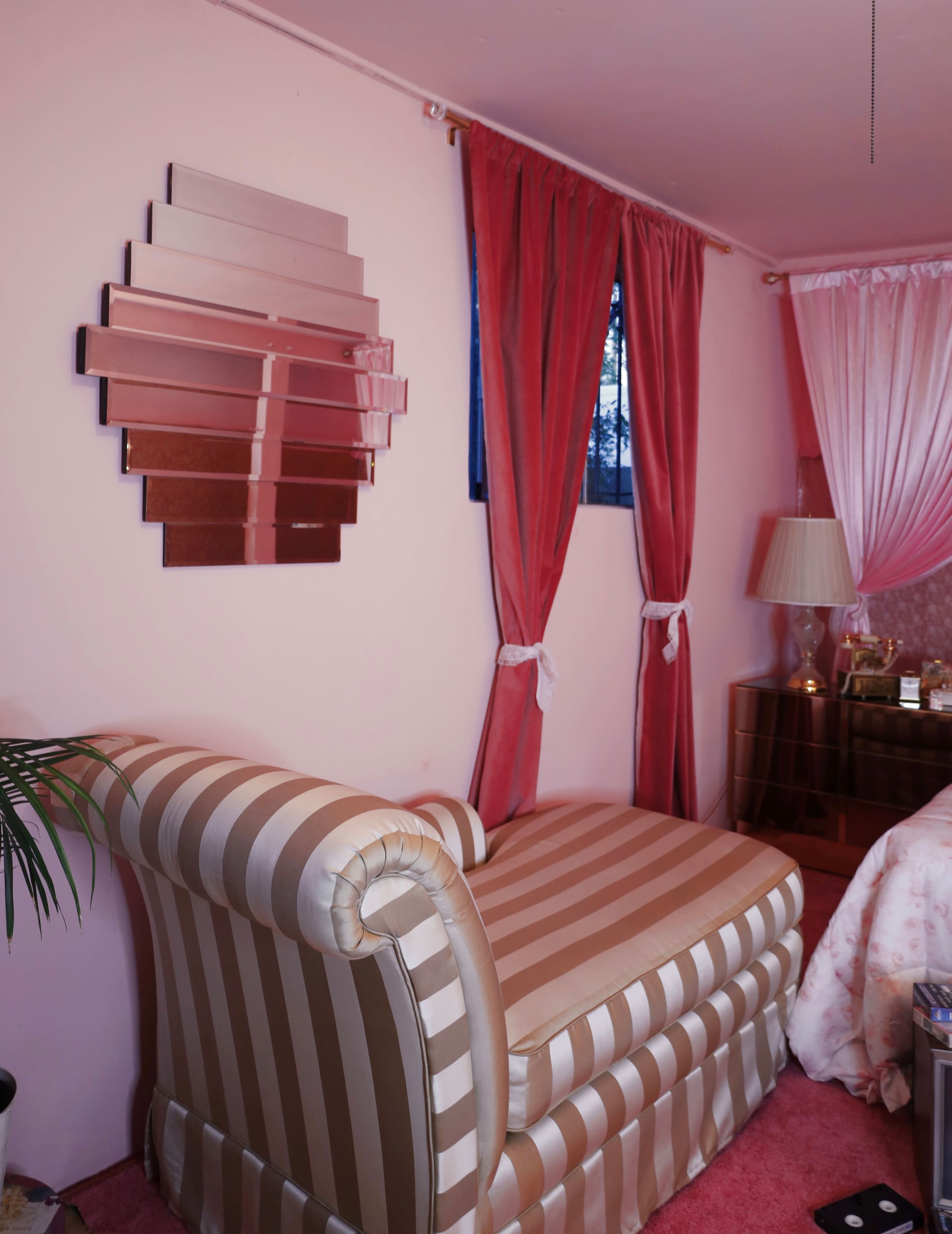 A pink room featuring a striped chaise lounge, a decorative mirror on the wall, and flowing curtains.