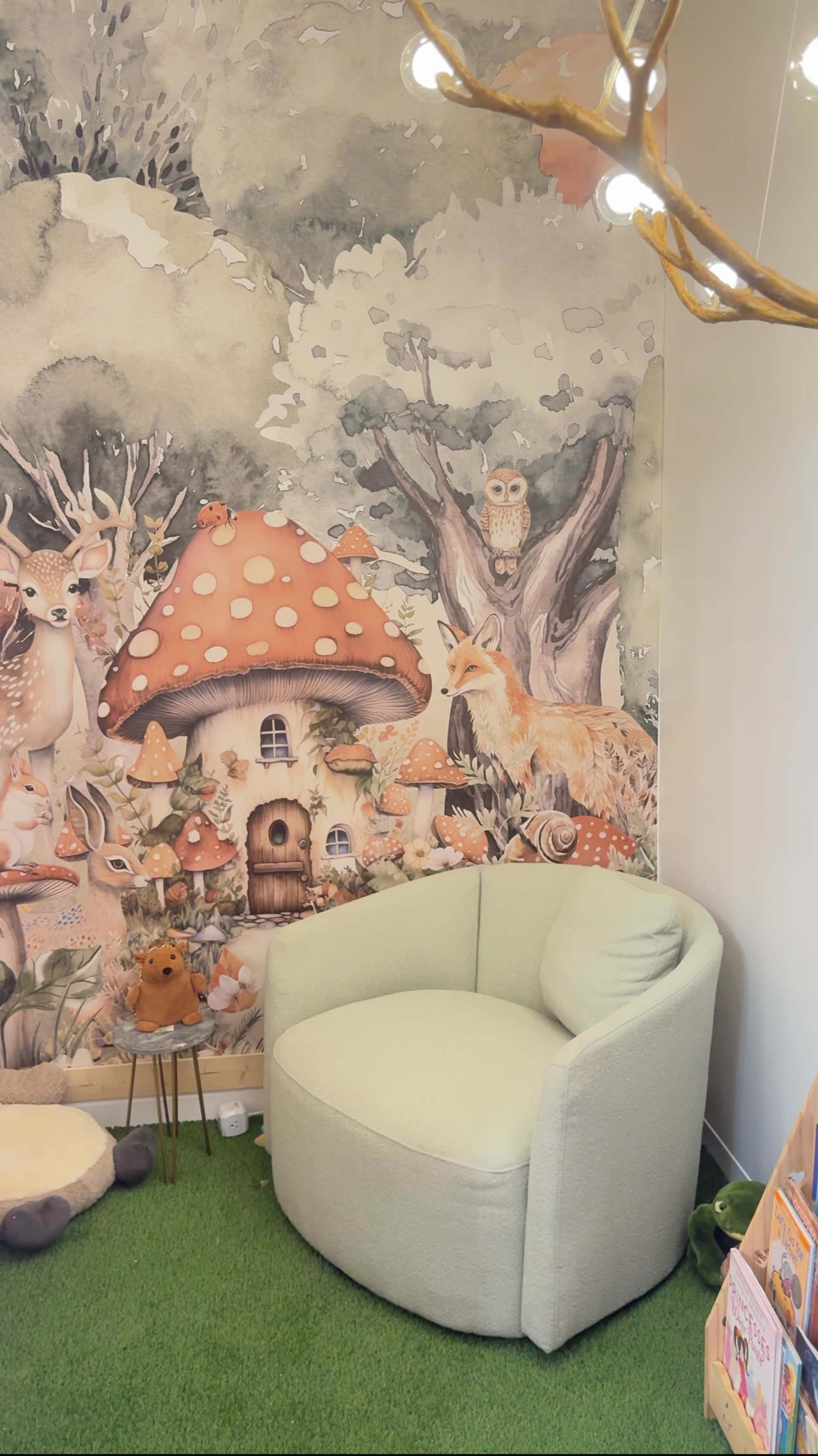 The image shows a cozy corner featuring a light green chair next to a whimsical wall mural of a forest scene with mushrooms, deer, and an owl.